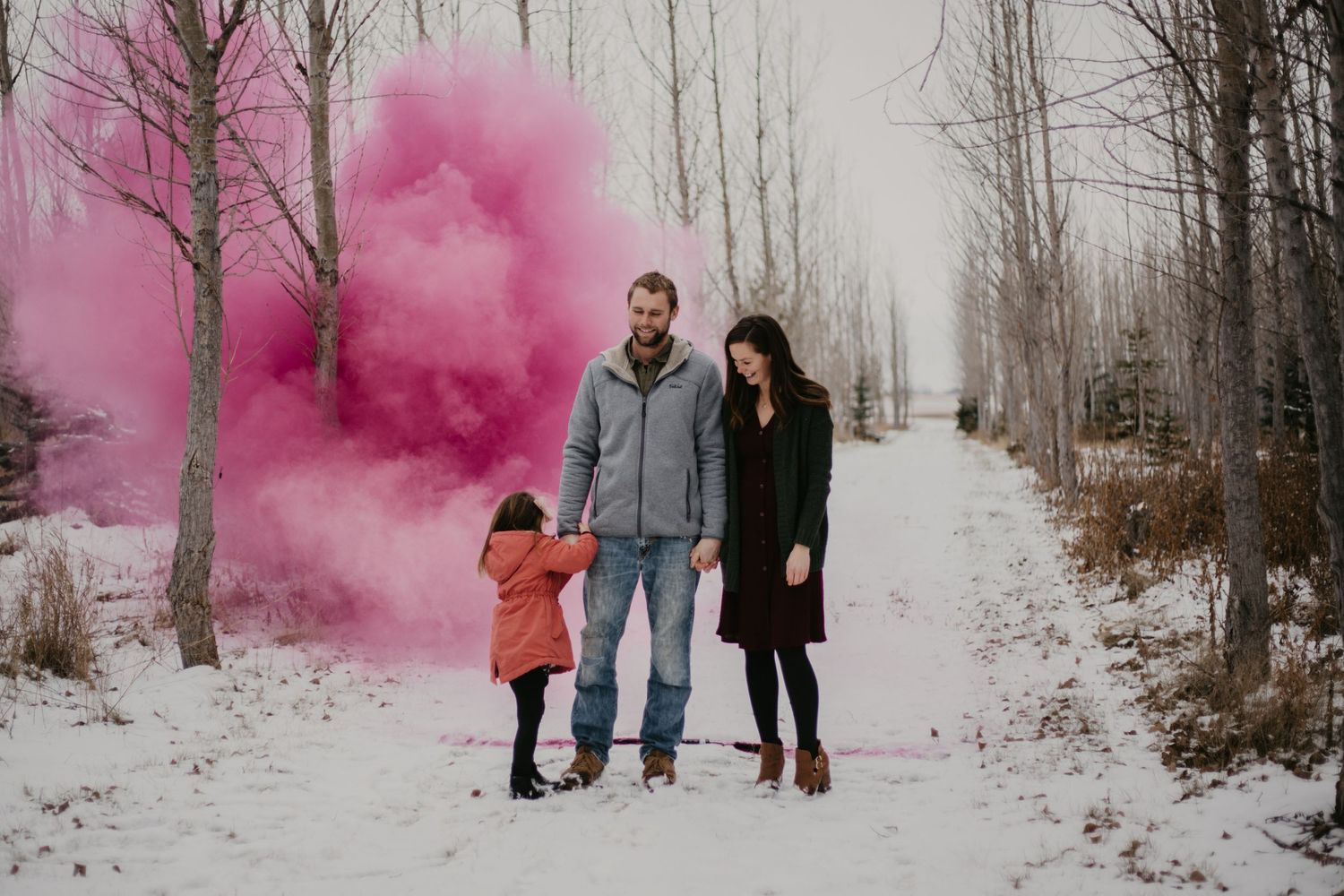 A family walks together on a snowy path with pink smoke creating a dreamy winter atmosphere.