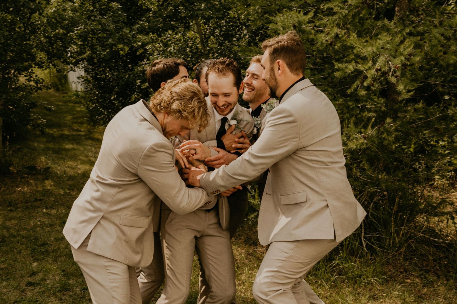 Groomsmen in tan suits share a playful group hug moment outdoors.