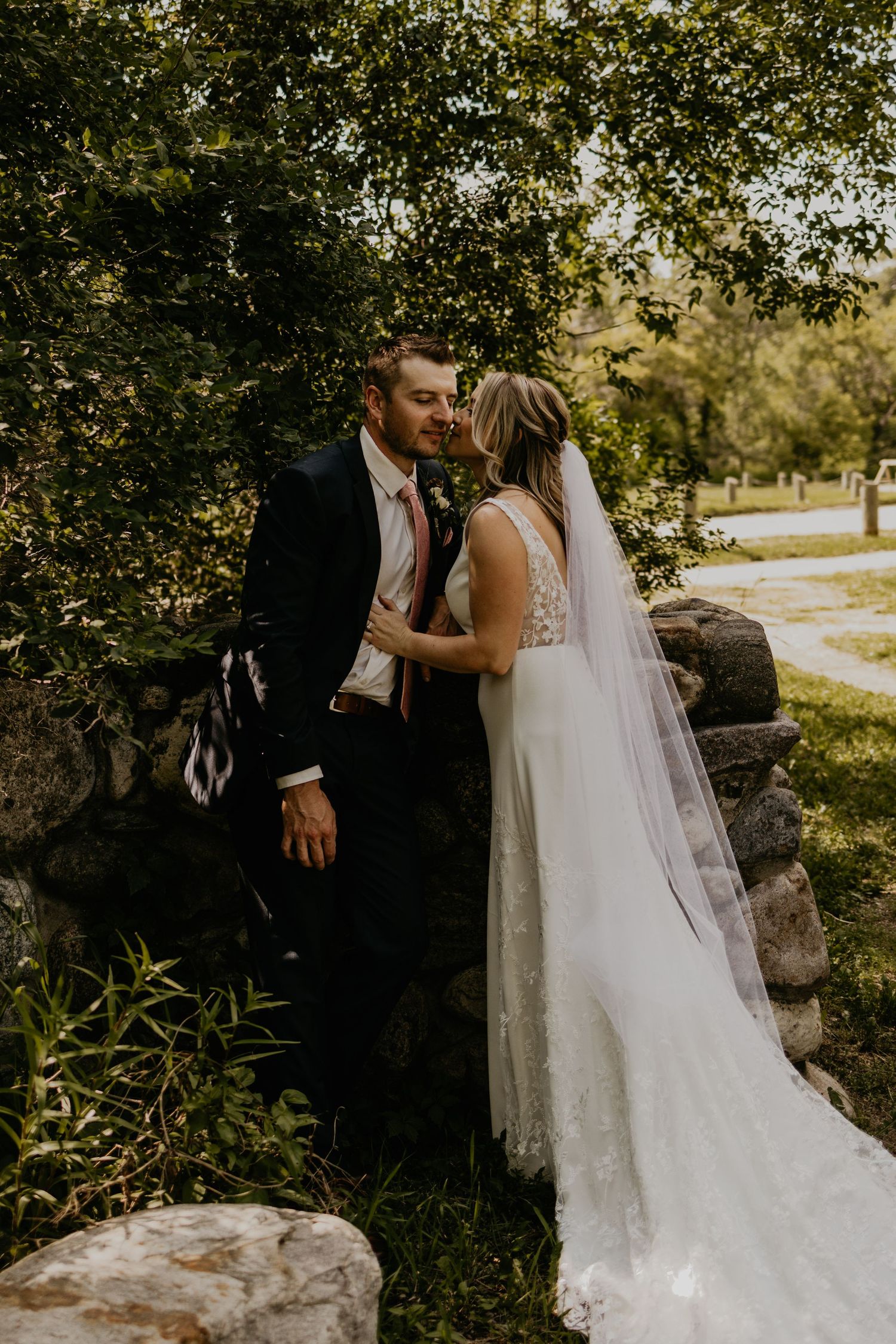 Candid wedding moment captured against stone wall with sunlit trees in background.