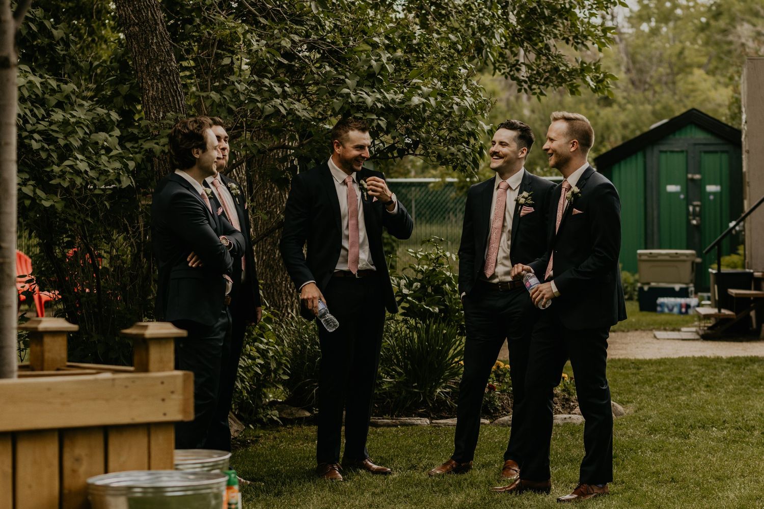 Groomsmen in dark suits share laughs during outdoor wedding gathering.