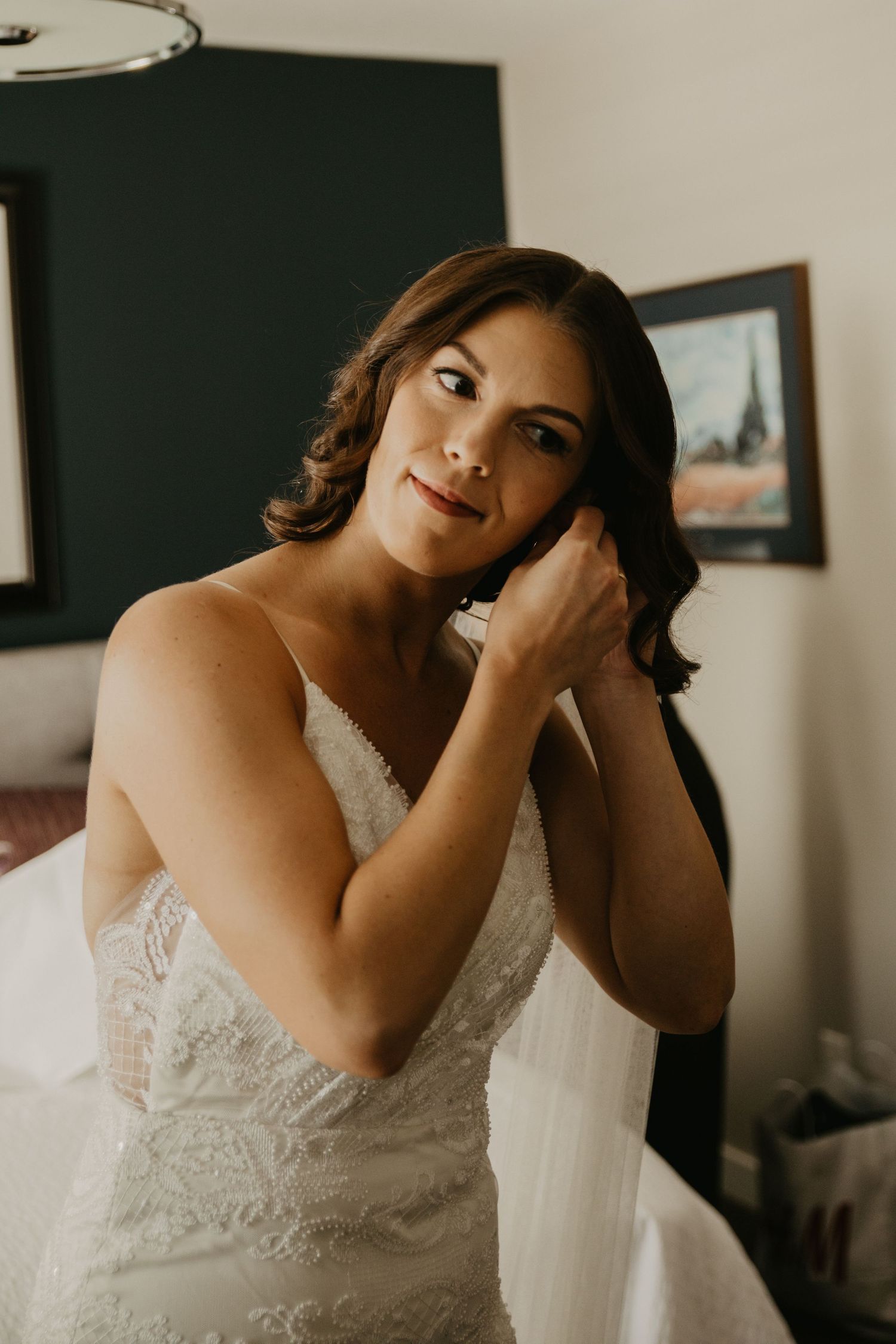 A bride wearing a lace wedding dress puts on earrings while getting ready.
