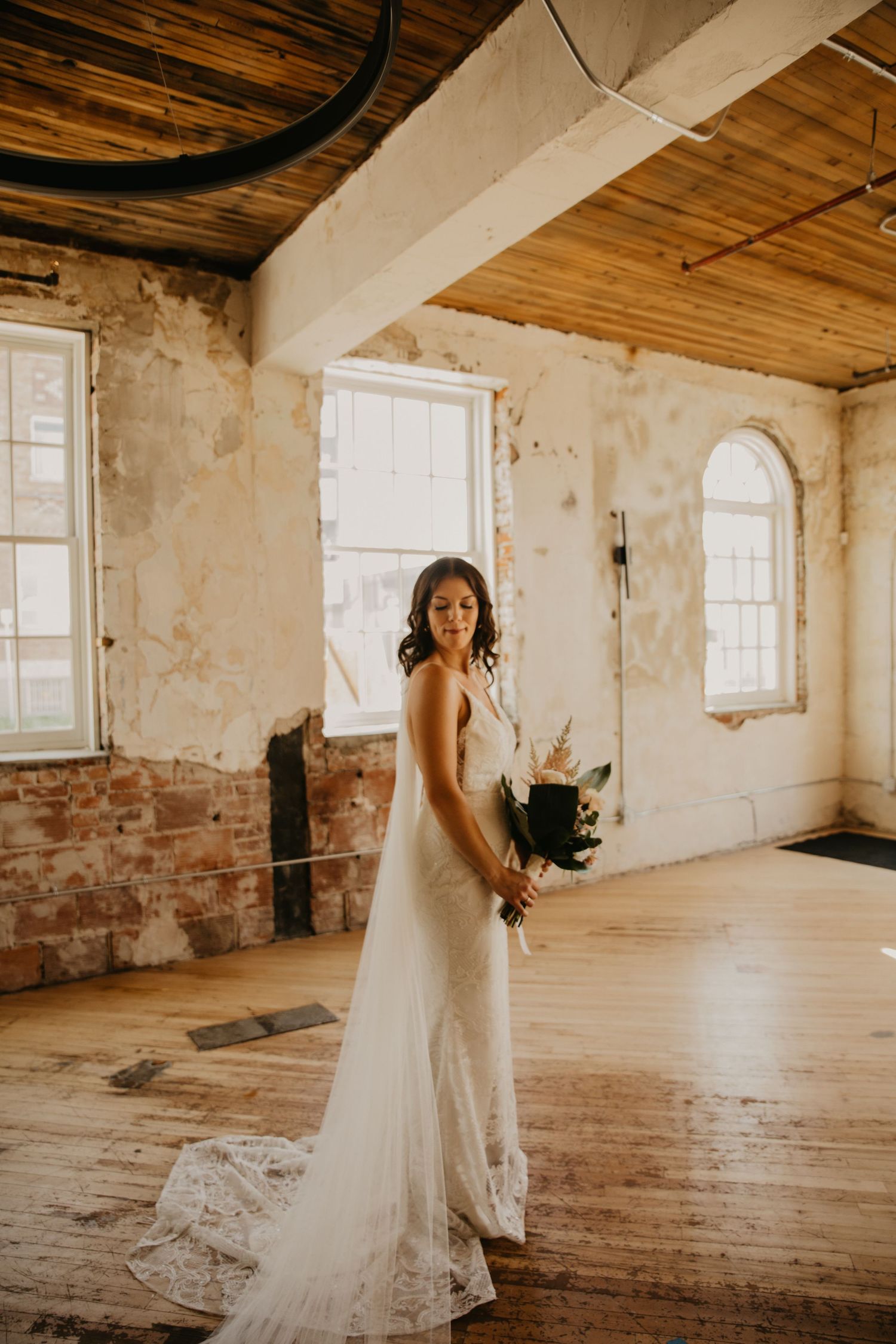 A bride in a flowing white lace gown poses in a rustic room with exposed brick walls and wooden floors.