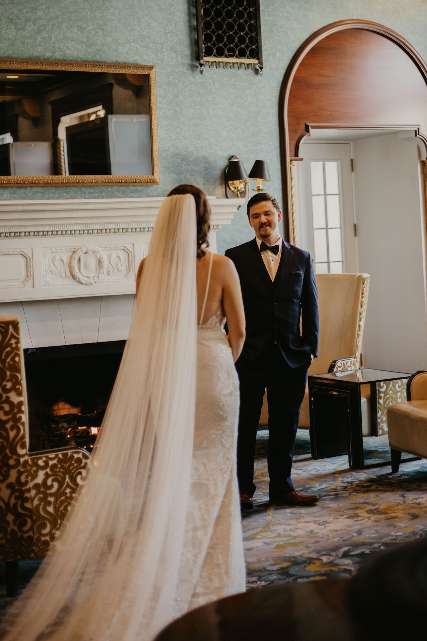A bride and groom share their first look in an elegant room with a white marble fireplace.