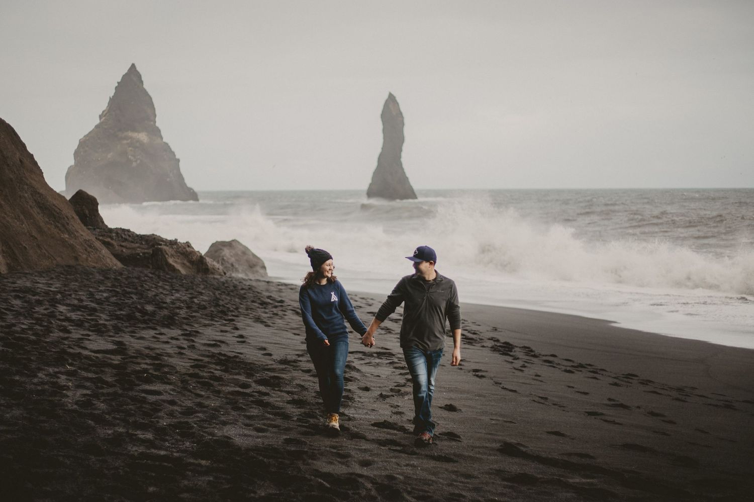 Two people strolling hand in hand on a moody black sand beach with rocky coastal formations.