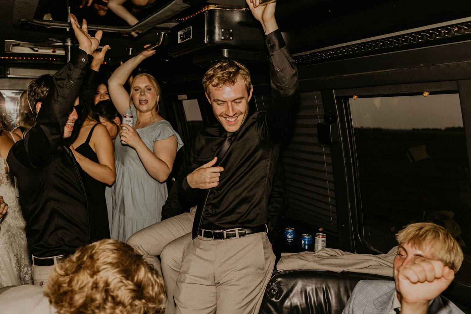 Wedding reception party guests dance enthusiastically with raised arms in a dimly lit venue.