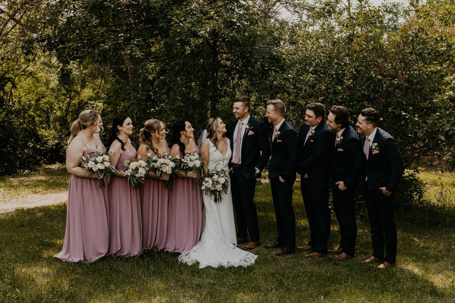 Wedding party in pink bridesmaid dresses and black suits poses together on a sunny lawn.