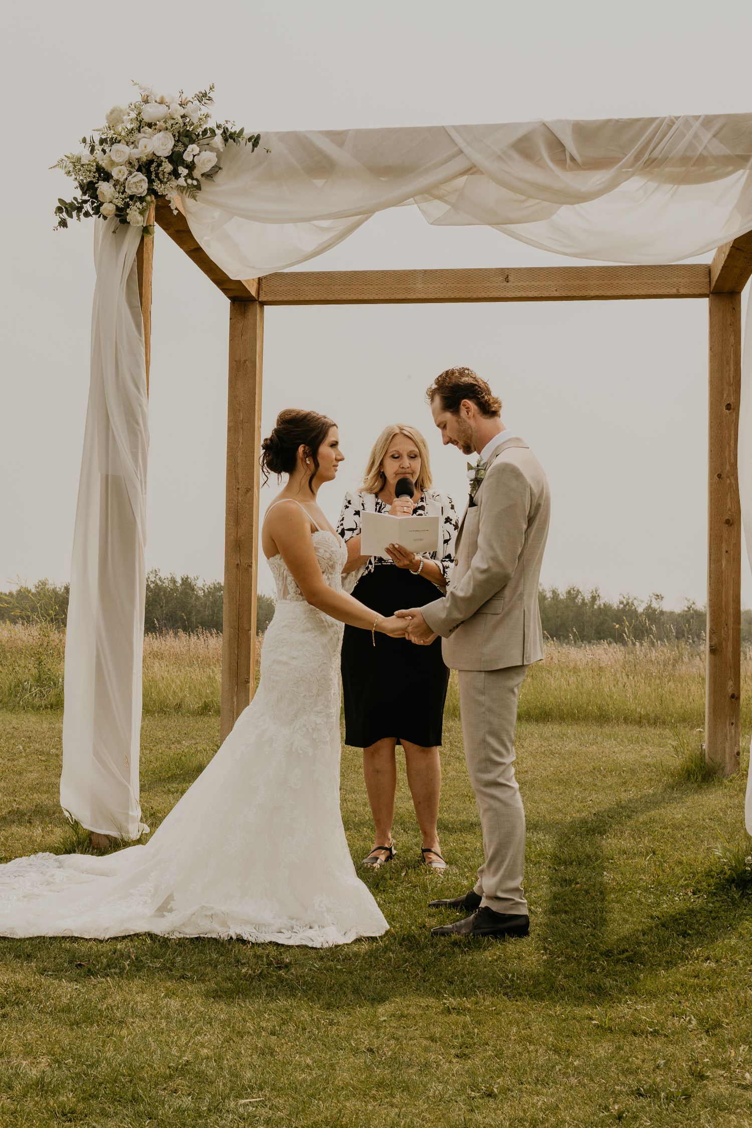 Outdoor wedding ceremony takes place under a wooden arch draped with white fabric and floral arrangements.