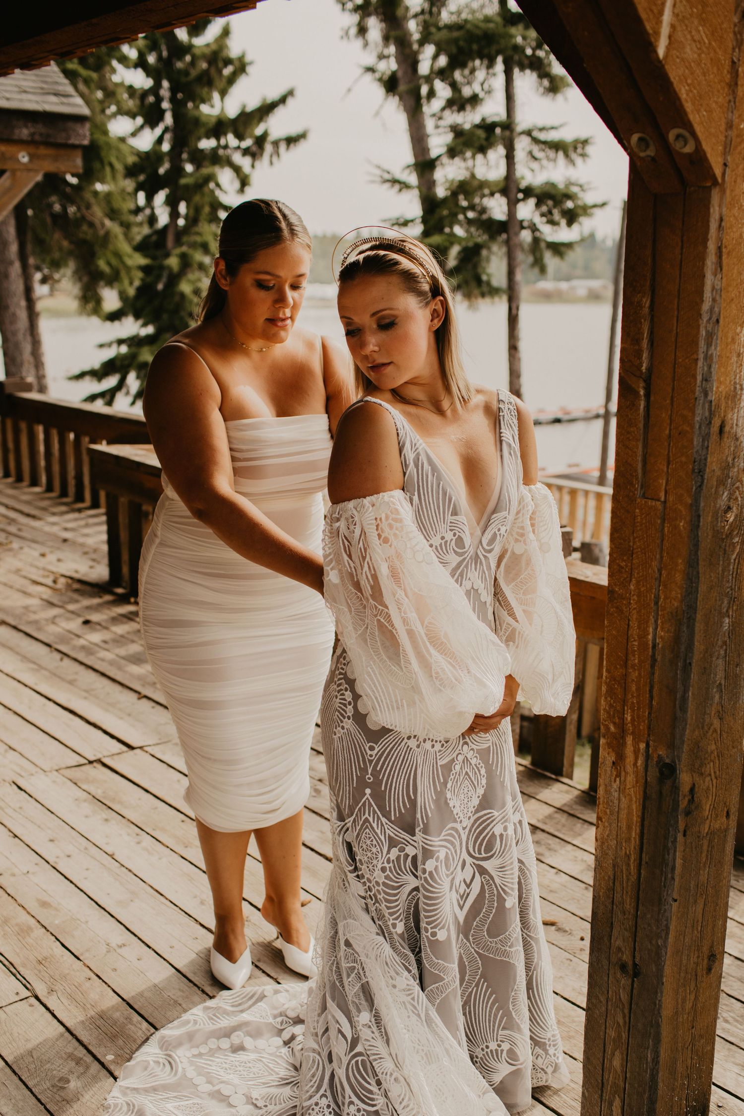 Two women in elegant dresses standing on rustic wooden deck overlooking lake.