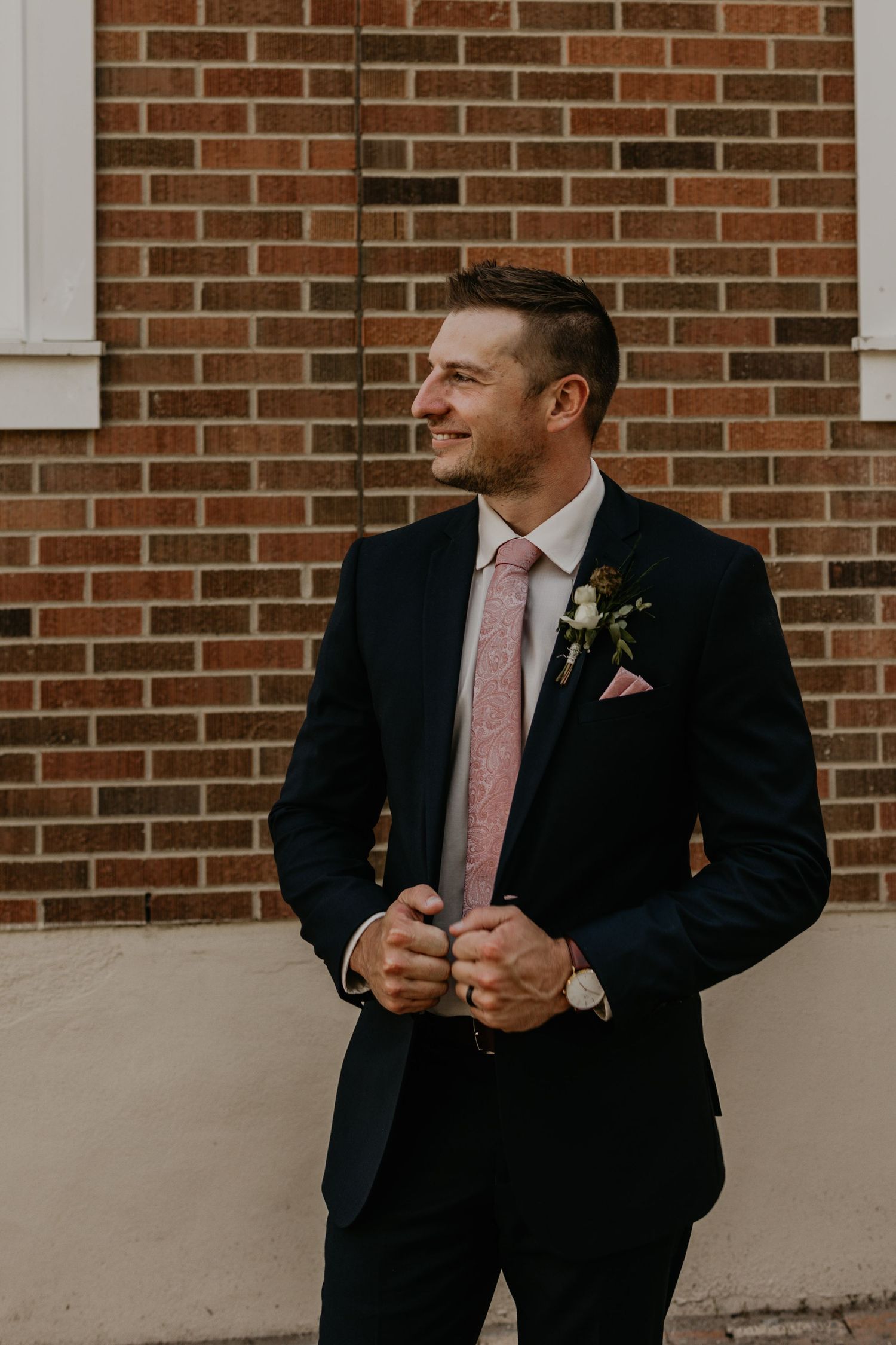 A stylish groom in a black suit and pink tie poses confidently against brick architecture.