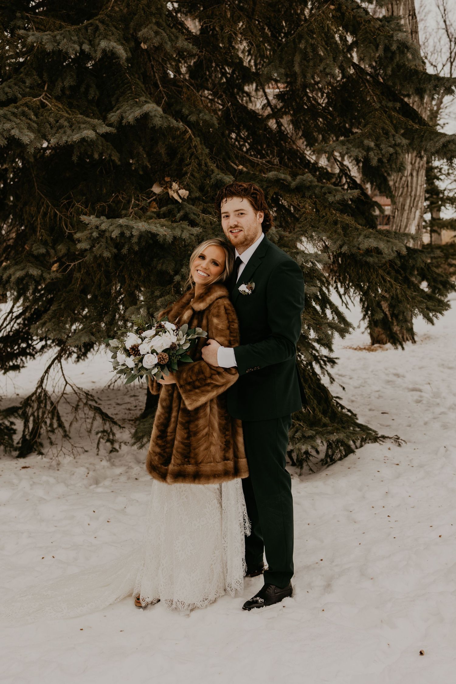 Newlywed couple embraces in snow under evergreen trees, bride in fur coat and groom in black suit.