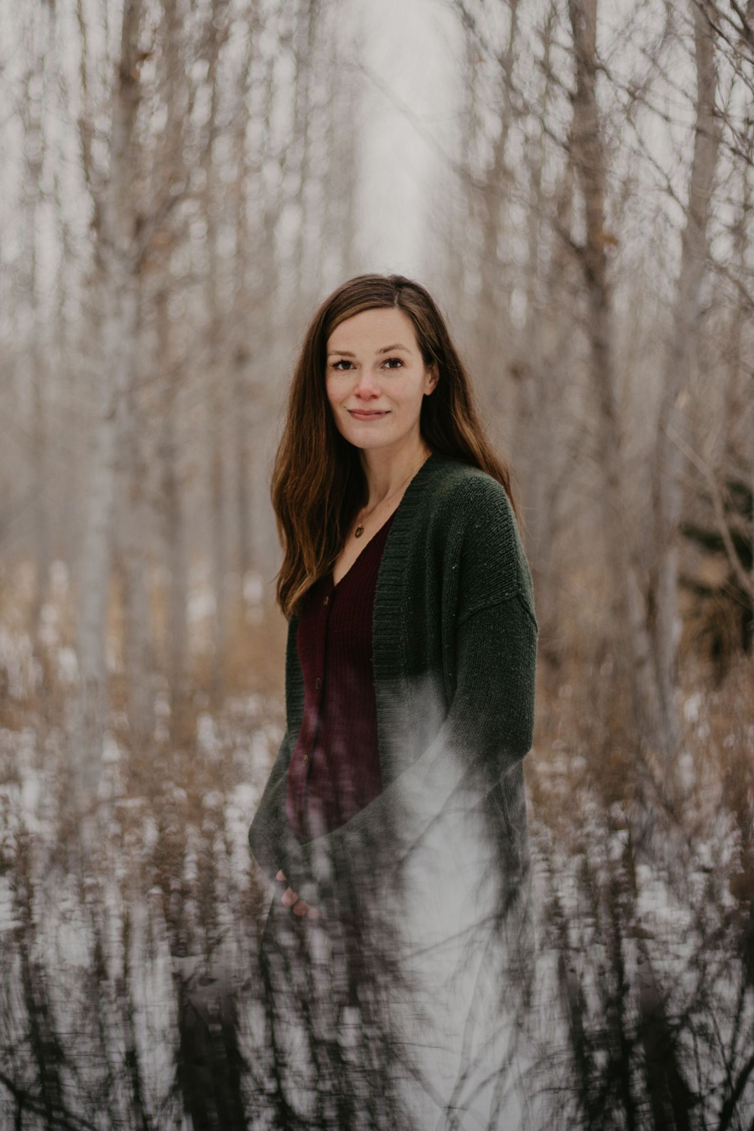 A person in a dark cardigan stands among winter trees creating a moody forest portrait.