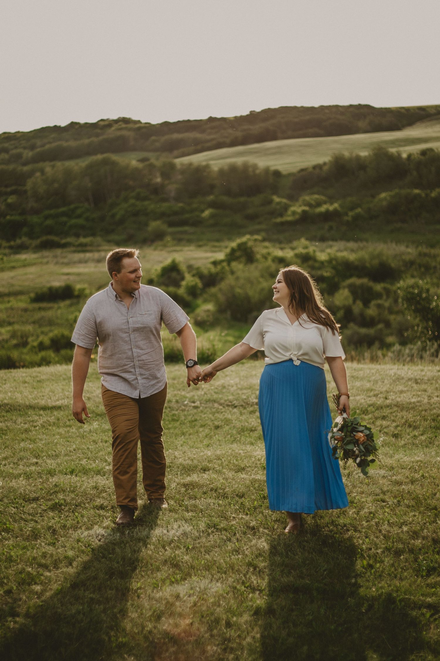 A couple walks hand in hand through a lush green hillside field at sunset.