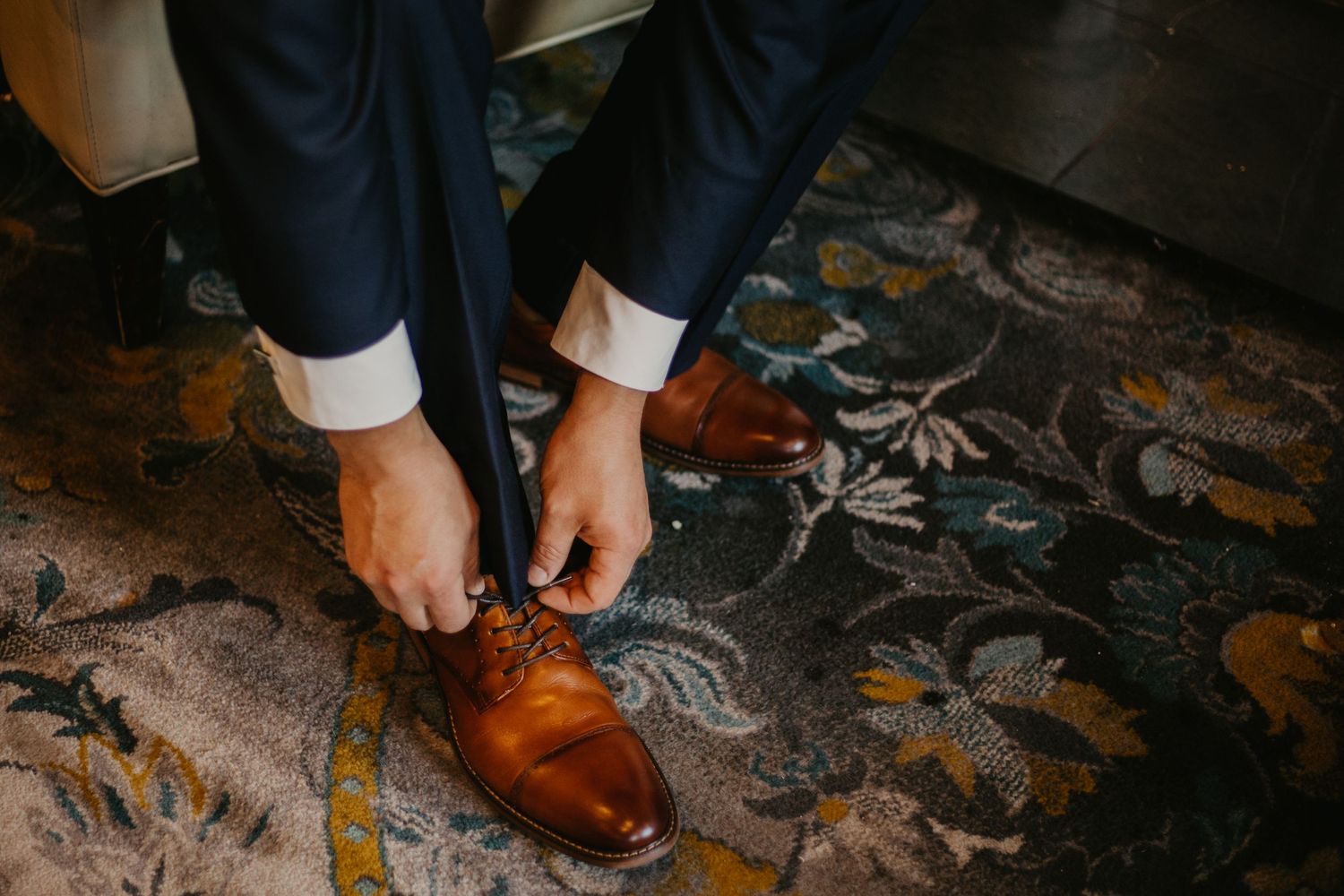 Close-up of brown leather dress shoes being laced up on patterned carpet.