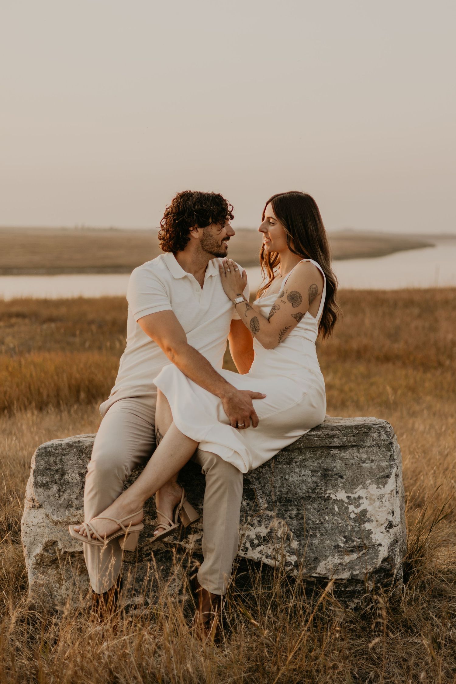A couple in light colored outfits sits intimately on a large rock in a golden field at sunset.