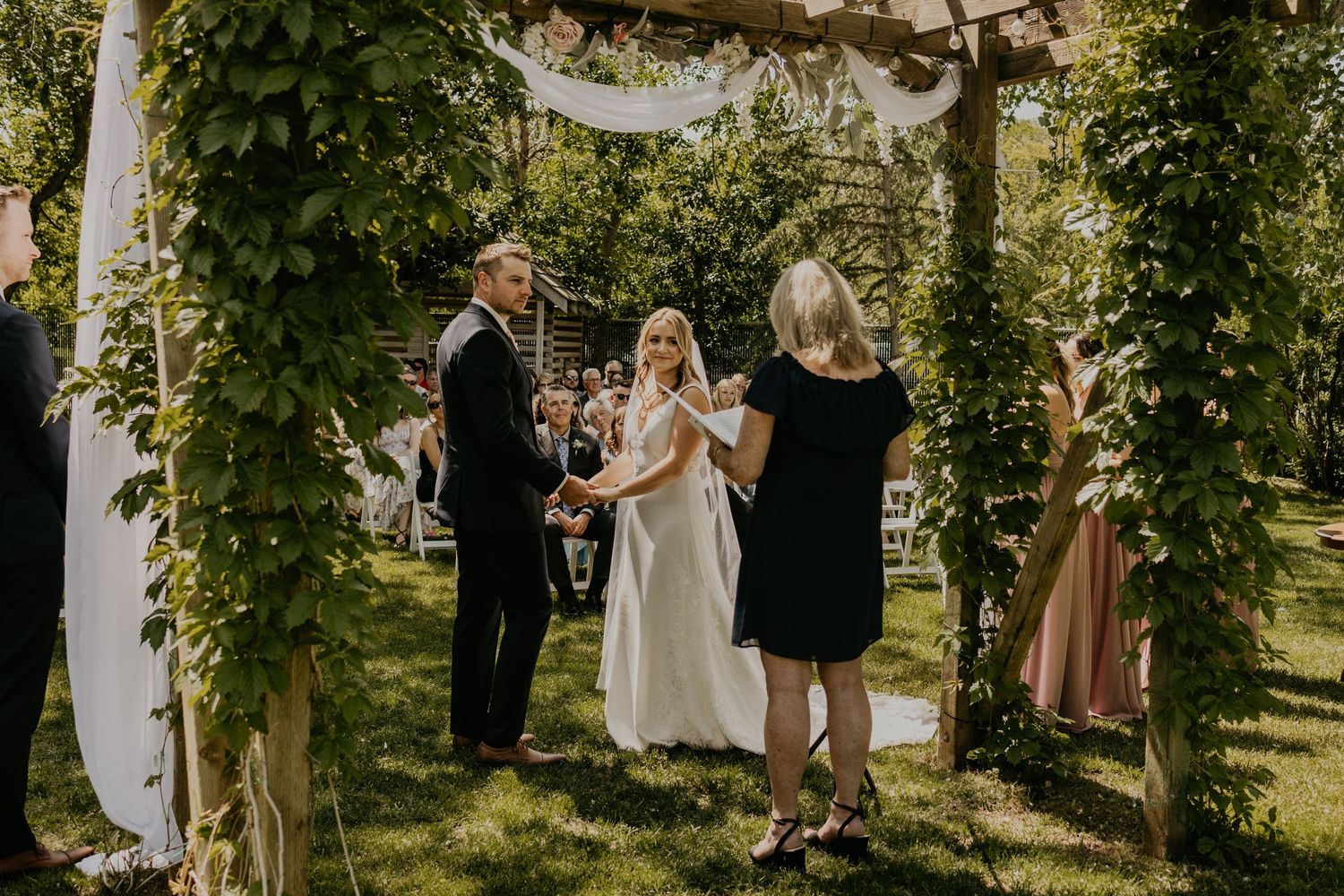 Wedding ceremony under a vine-covered arbor with white fabric draping in a garden setting.