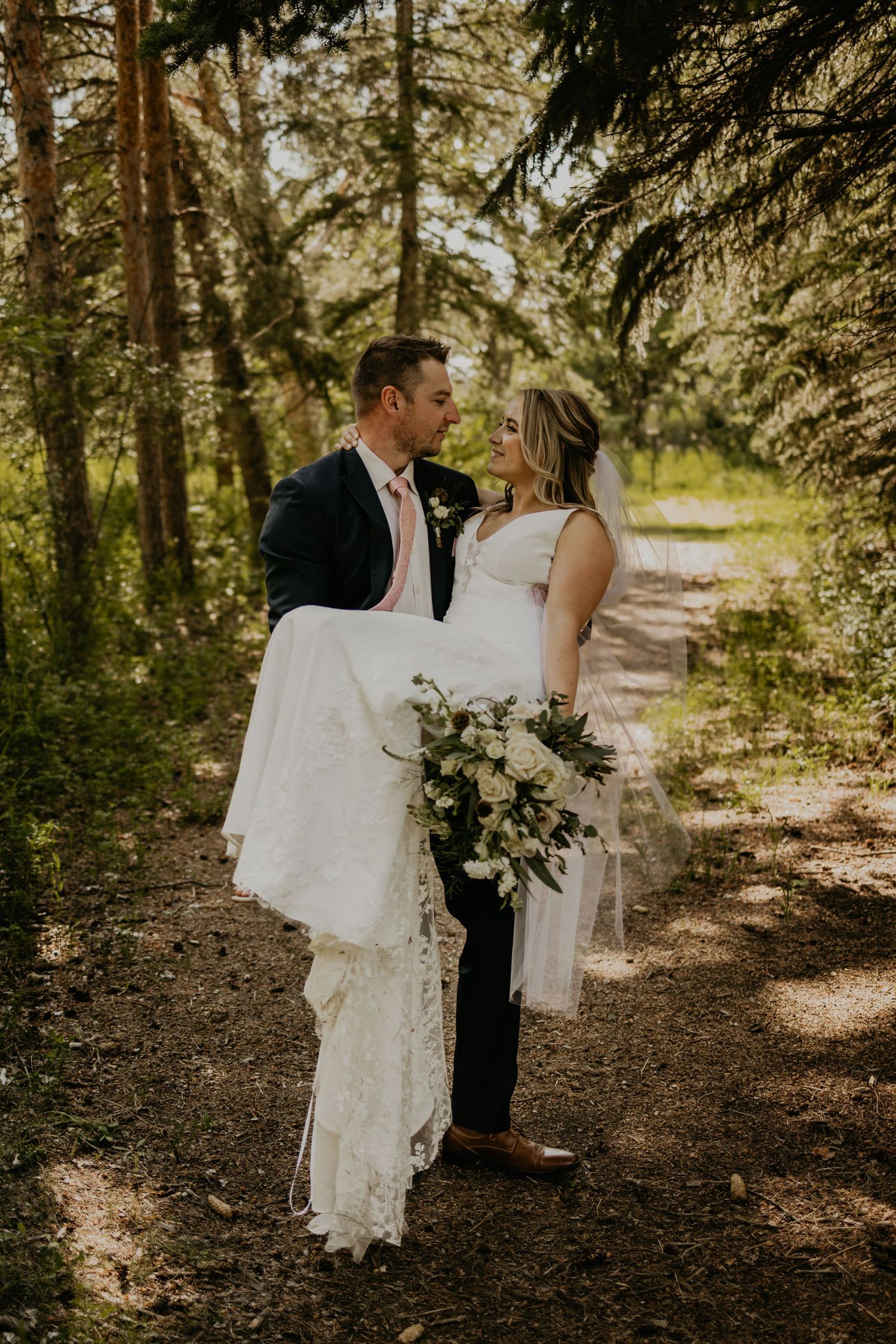 A groom carries his bride while standing on a forest trail surrounded by pine trees.