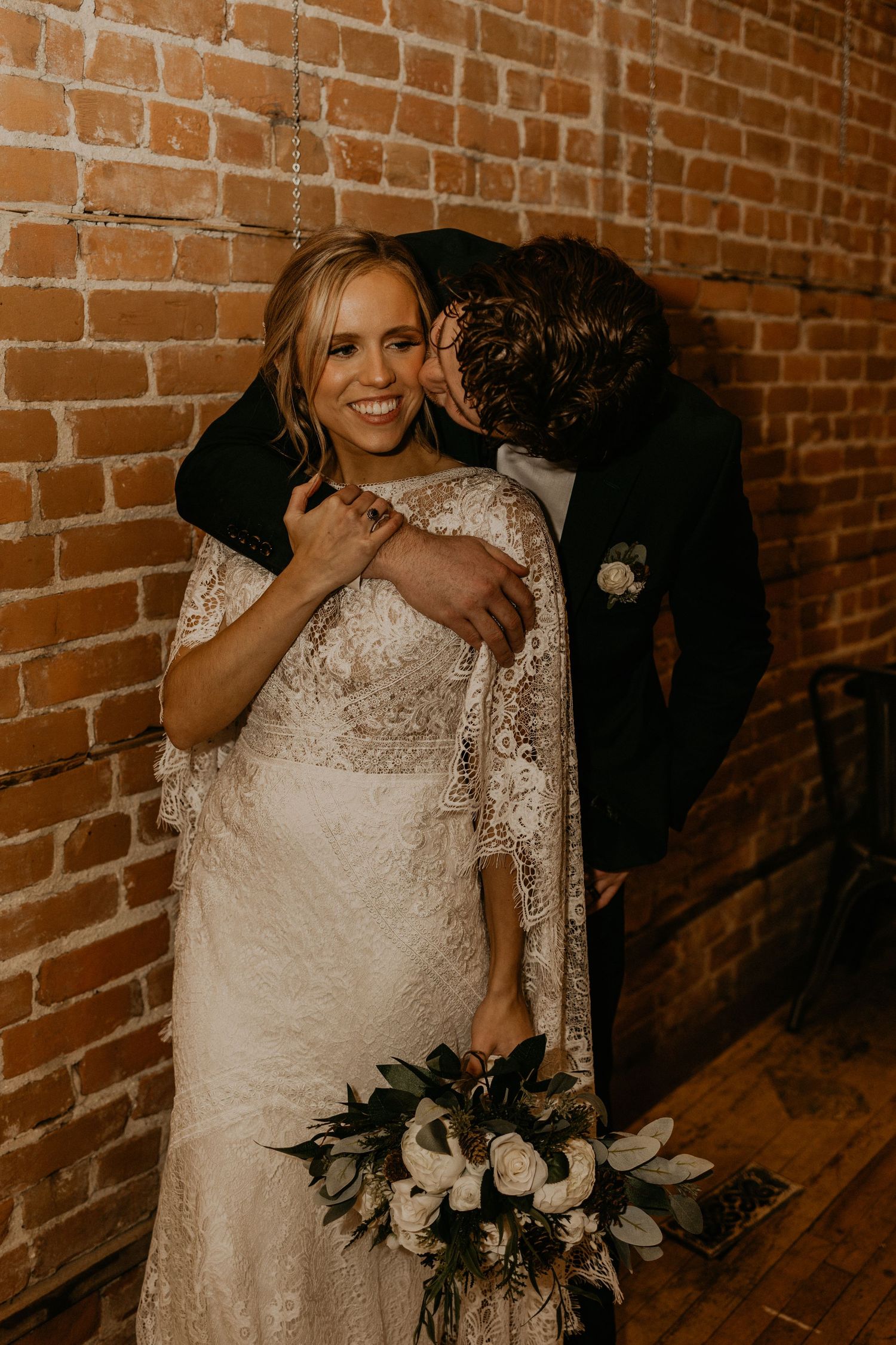 Candid wedding moment captured against brick wall with white floral bouquet.