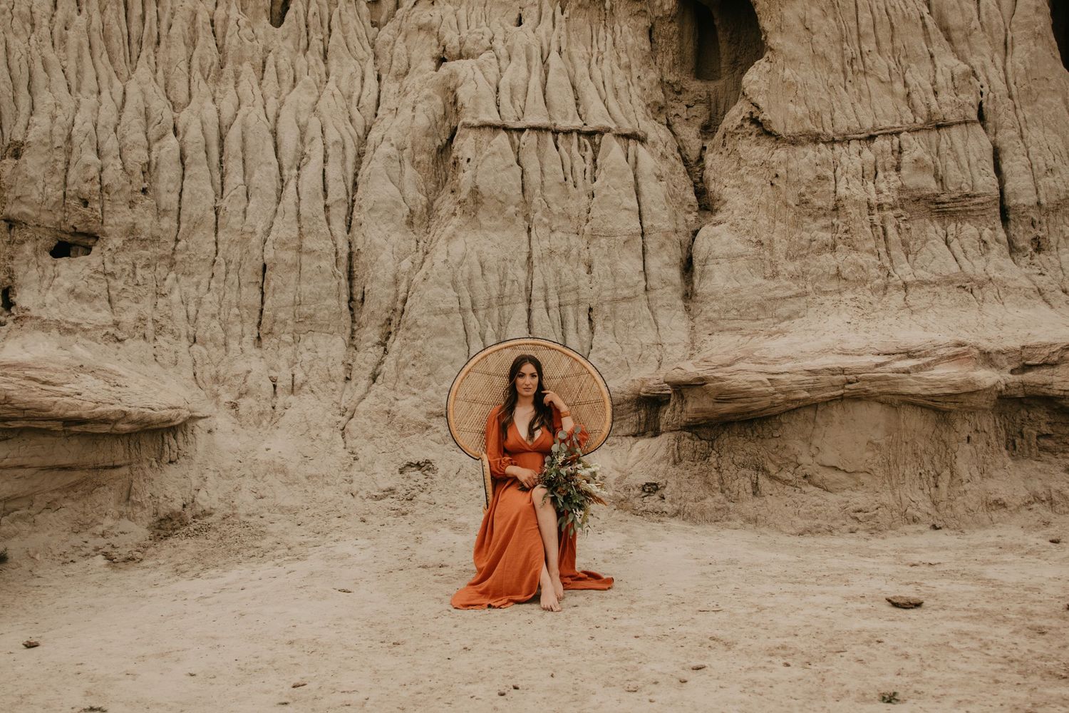 Figure in flowing orange dress poses with wicker chair against weathered clay cliffs.