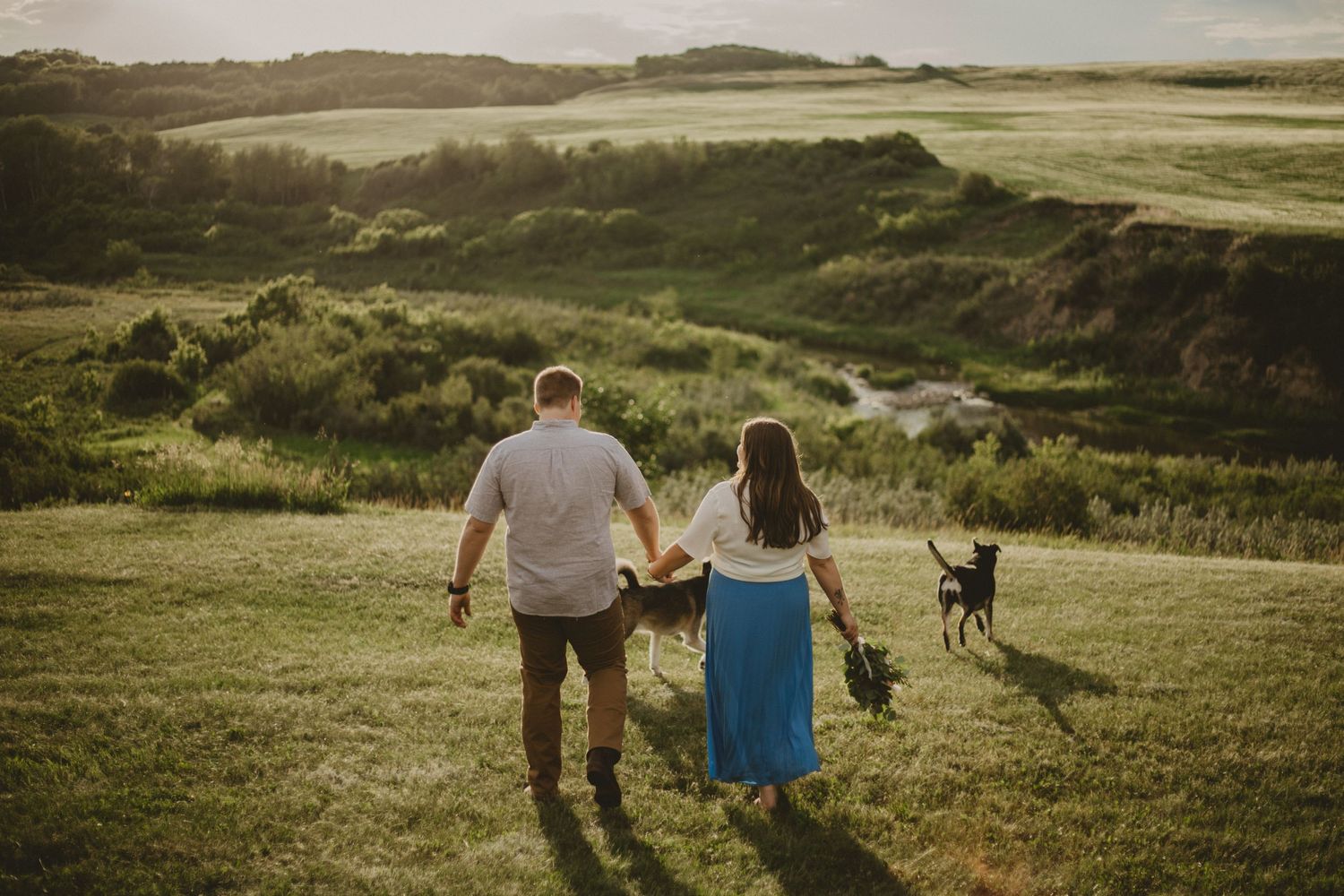 A couple walks along a grassy hilltop path with their dog at sunset.