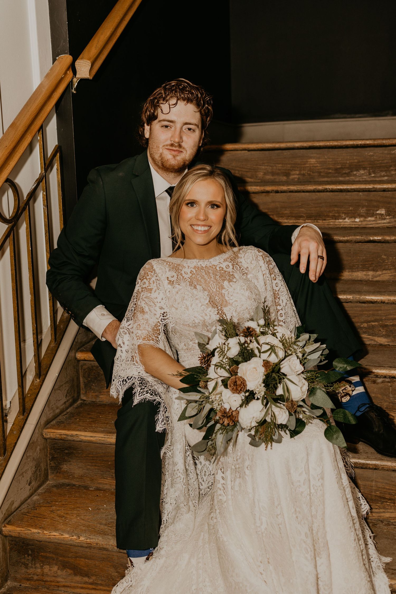Wedding couple poses on wooden staircase with elegant bouquet and lace dress details.