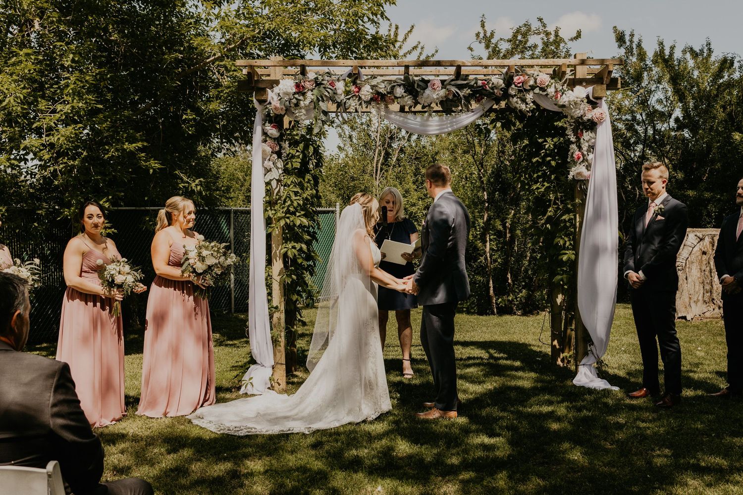 Wedding party gathered under a floral arch during an outdoor ceremony with bridesmaids in pink dresses.