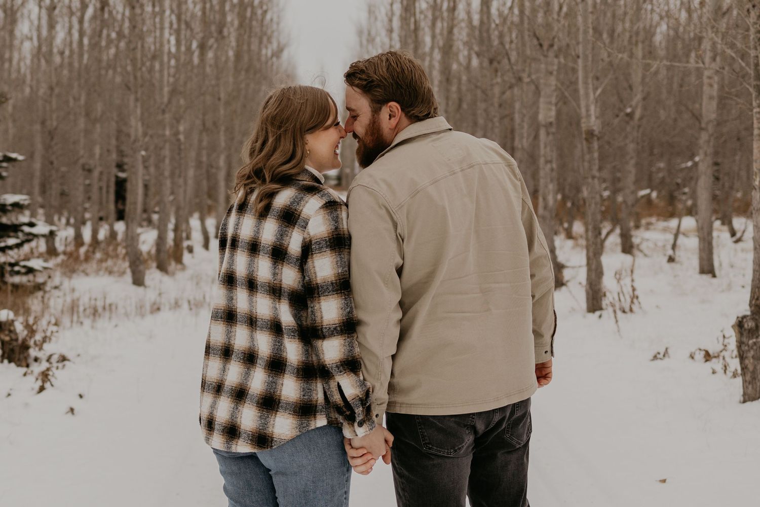 Couple walking hand in hand through snowy forest path wearing cozy winter clothing.