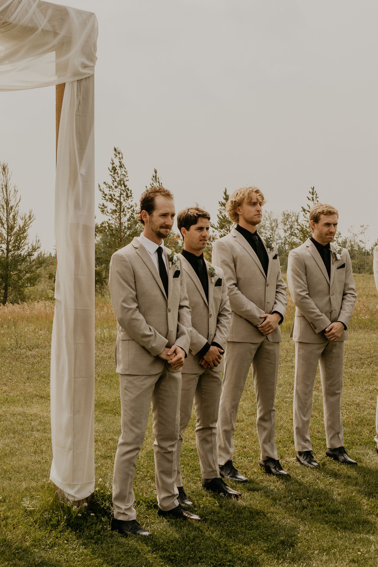 Groomsmen in matching beige suits stand in a line under a white wedding arch during an outdoor ceremony.