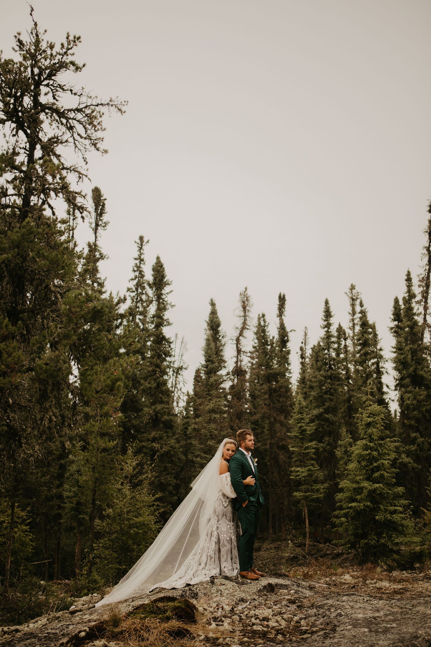 A couple in wedding attire stands on a rocky outcrop surrounded by tall evergreen trees.