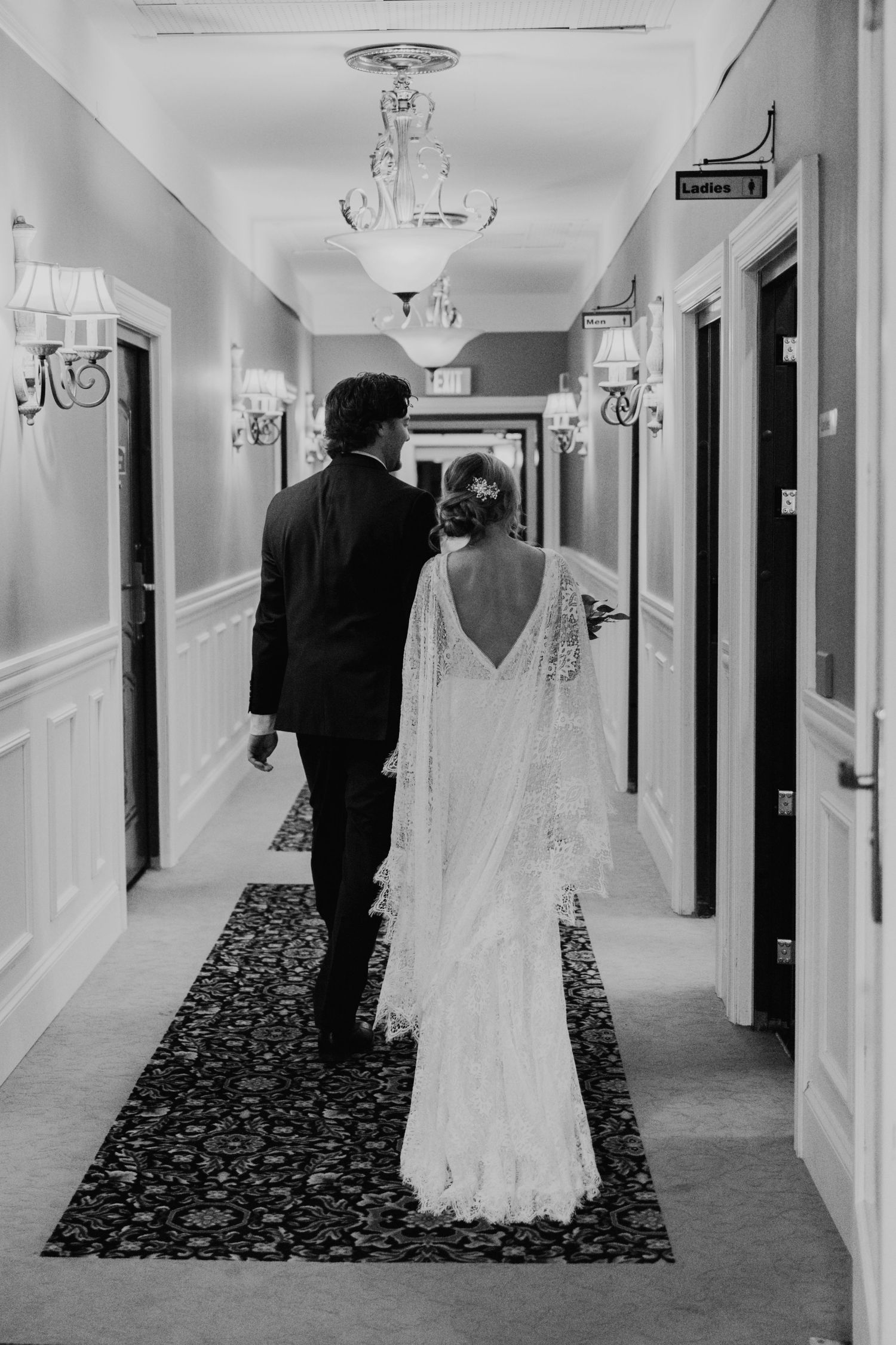 A black and white wedding photo capturing a couple walking down an elegant hallway with chandeliers.
