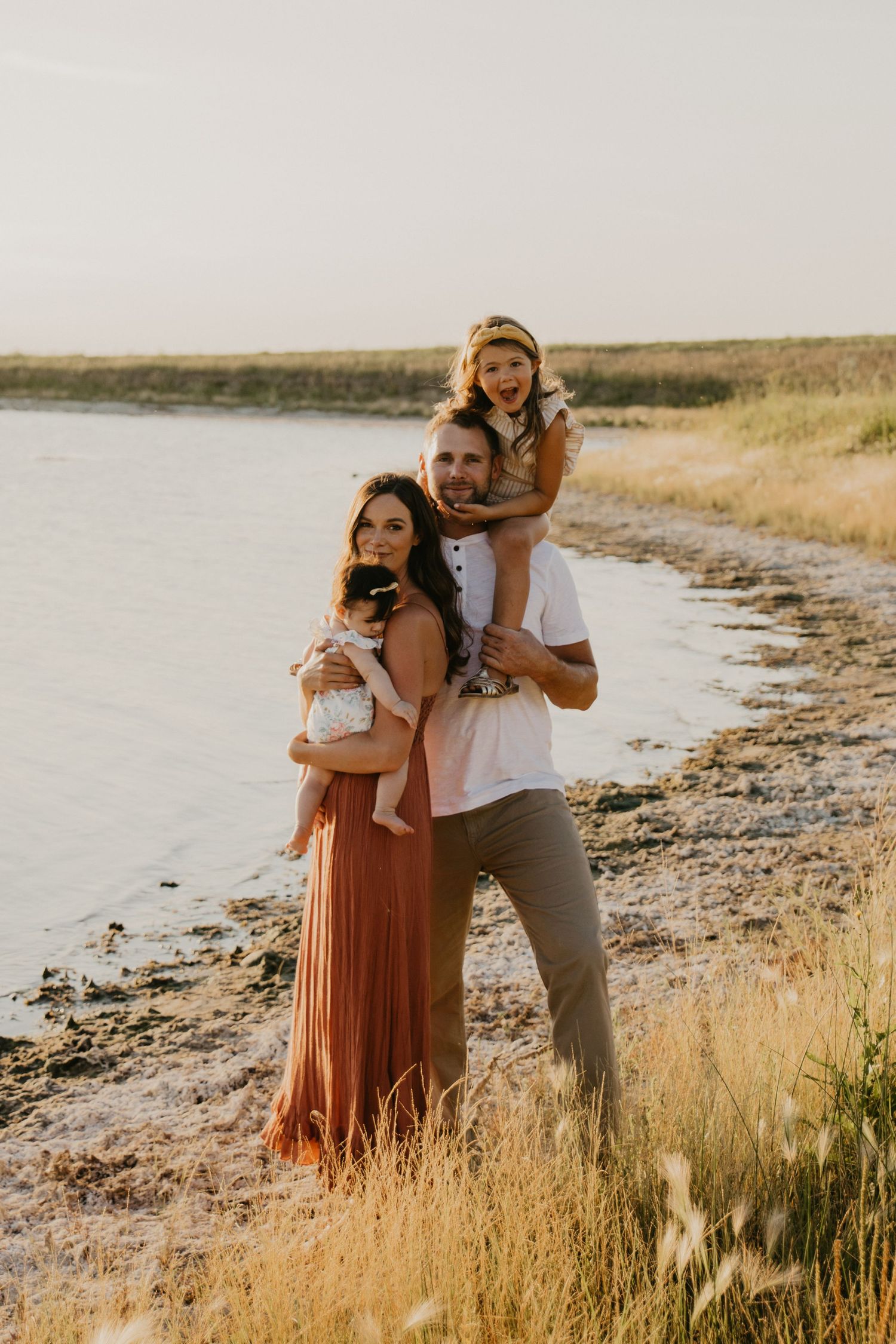 Family portrait at waterside with golden grass and natural lighting at sunset.