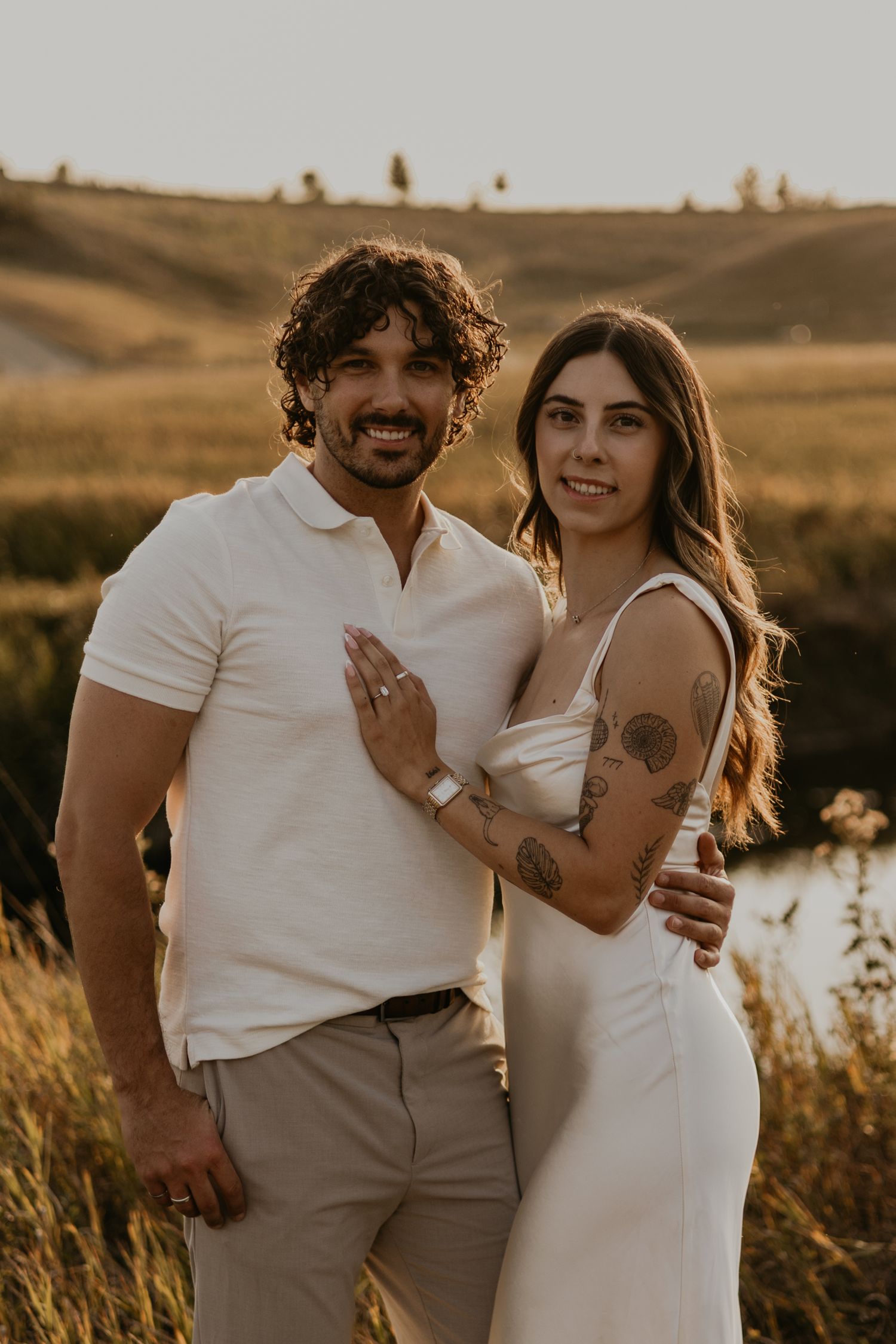 Couple embraces in a golden field during sunset while wearing casual white and beige outfits.