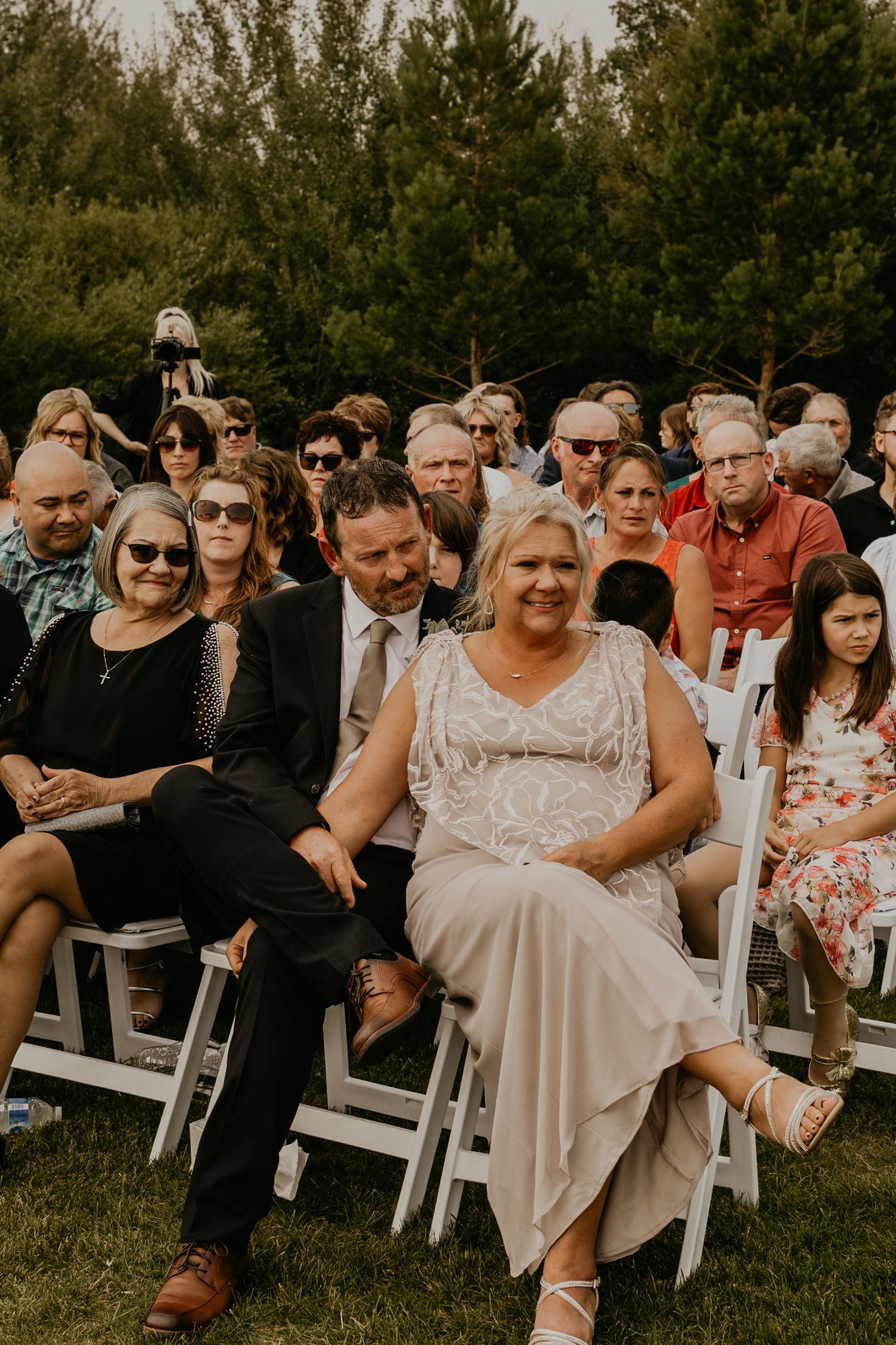 Wedding guests seated in white chairs watch ceremony in outdoor garden setting.