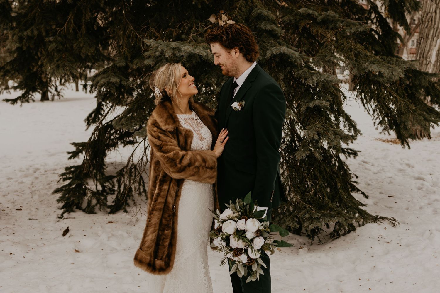 Winter wedding couple shares romantic moment beneath evergreen trees in snow.