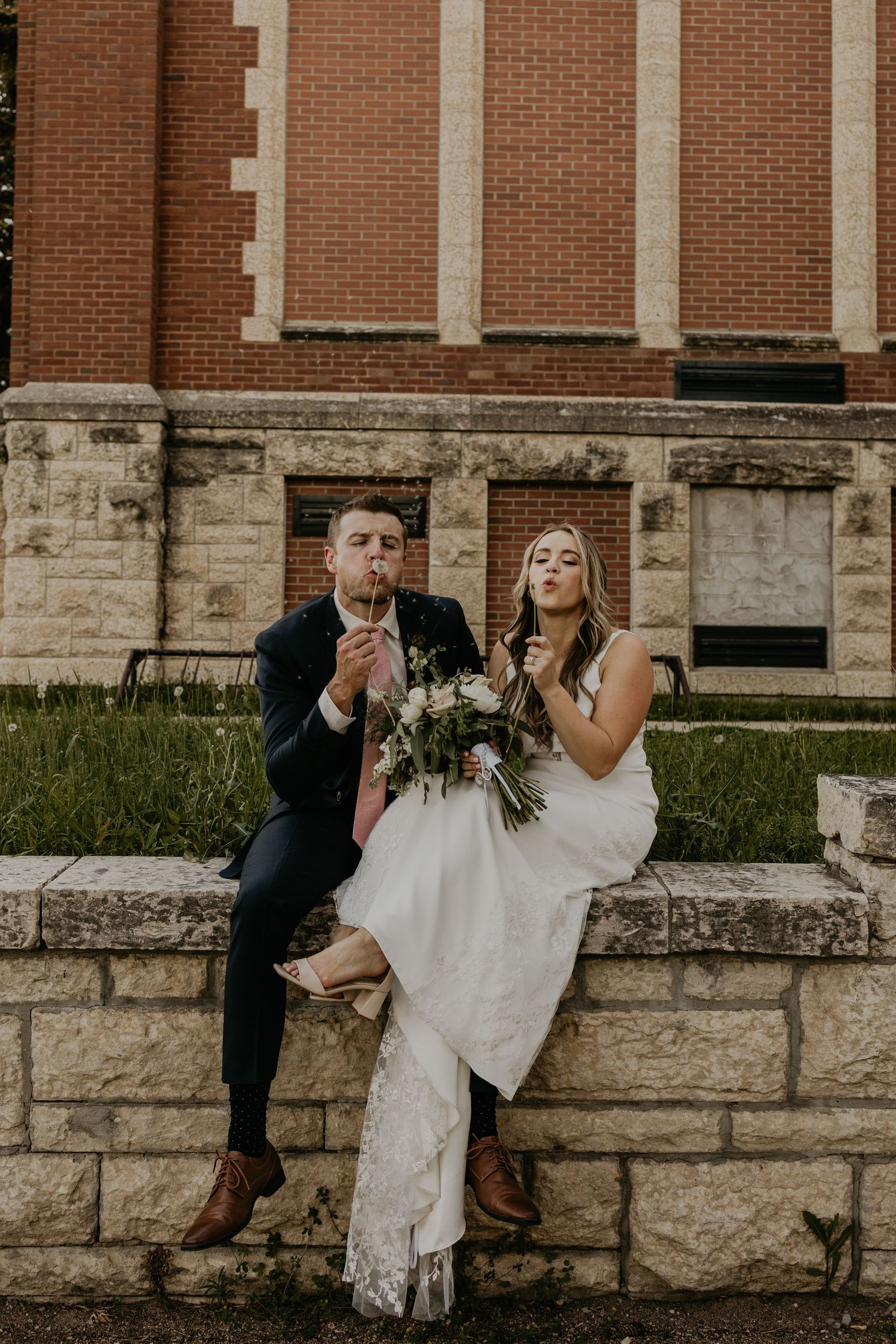 Newlyweds share a playful moment while sitting on stone steps of a brick building.