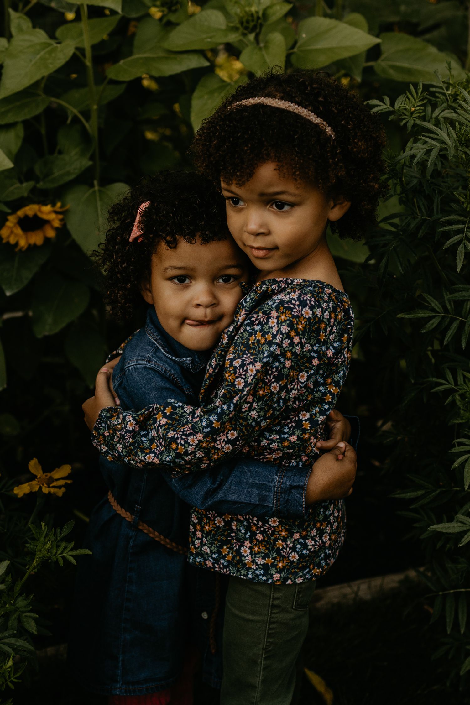 Two young children in floral and denim outfits hugging in front of sunflowers.