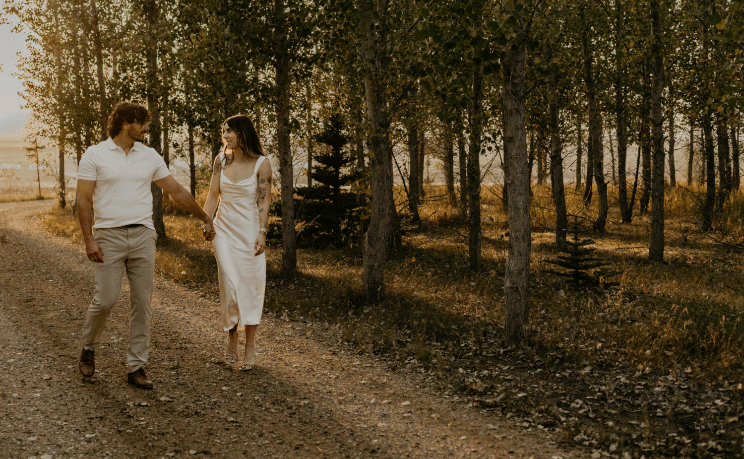Two people in light-colored clothing walk hand in hand down a dusty path through a golden-lit forest at sunset.