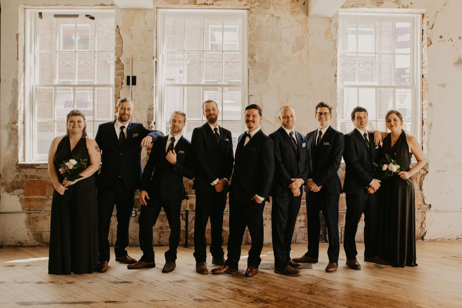 Wedding party members in black suits and dresses stand together in a bright rustic venue.