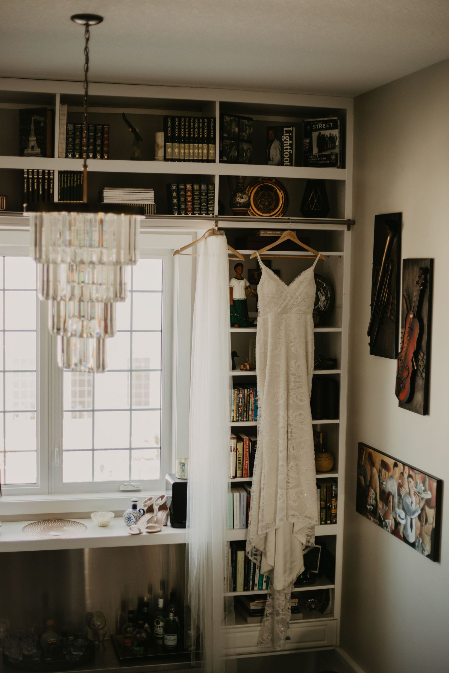 An elegant white wedding gown hangs in front of a built-in bookshelf with a crystal chandelier.