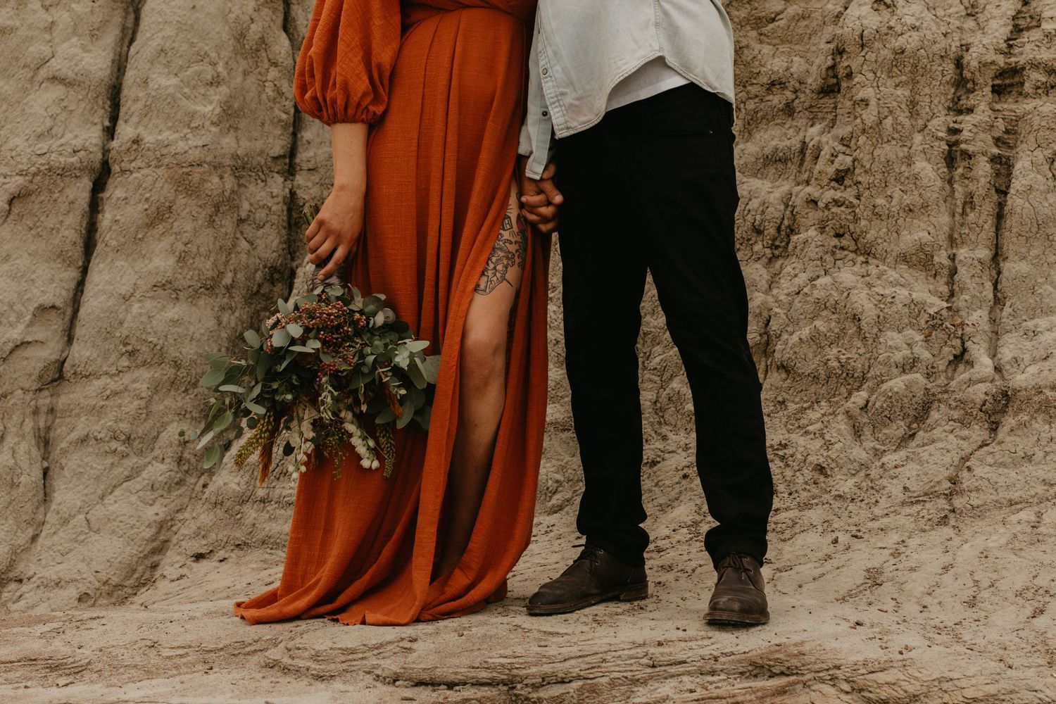 Lower body shot of a couple standing against clay cliffs with a flowing rust-colored dress and bouquet.
