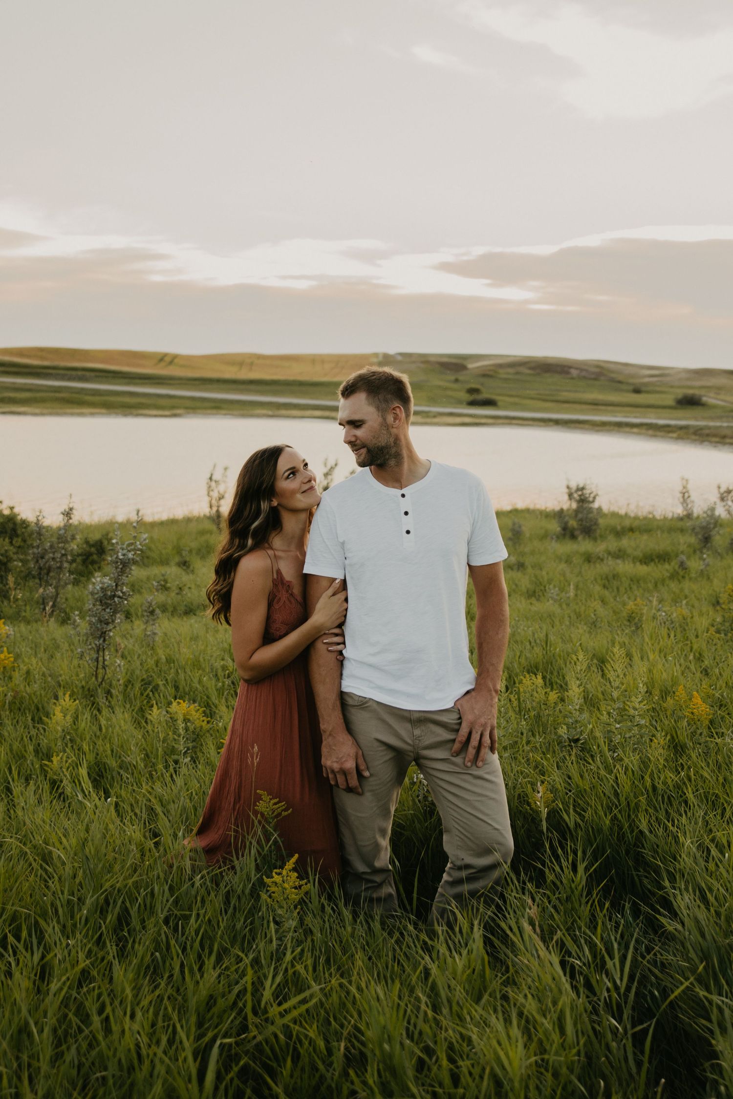 Couple standing in tall grass field beside lake during sunset.