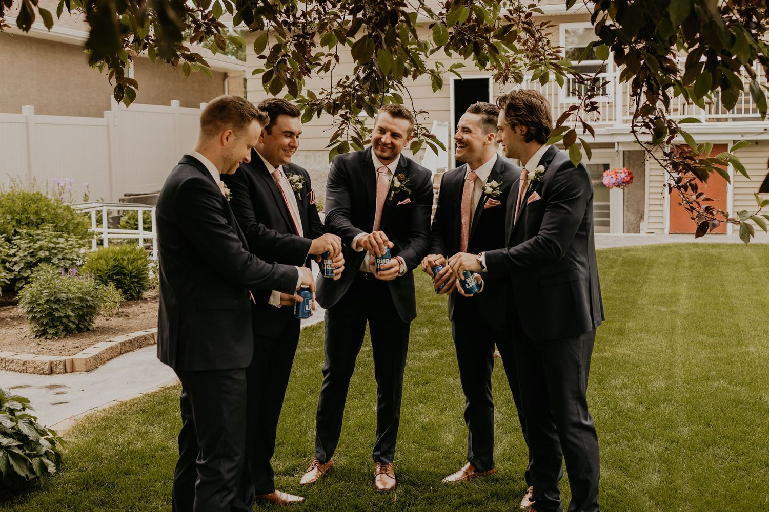 Group of groomsmen gathered in circle on lawn sharing pre-wedding drinks.