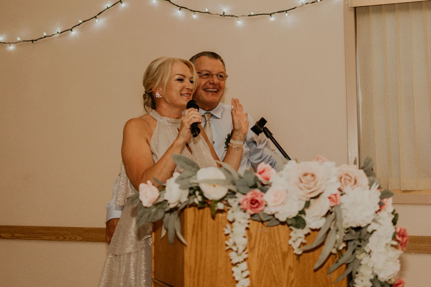 Wedding reception speeches at a podium decorated with pink and white flowers and string lights.