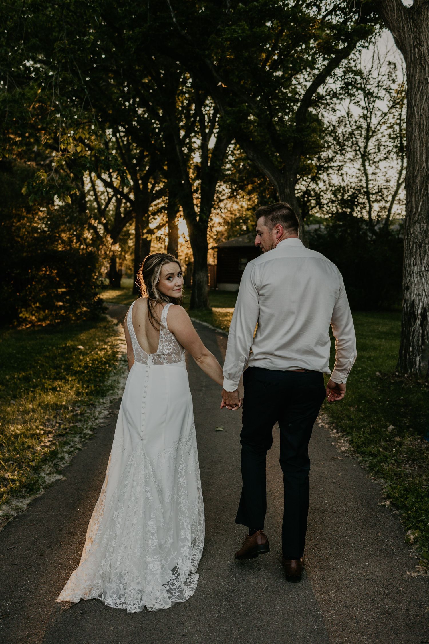 A couple walking hand in hand on a sunset path wearing wedding attire.