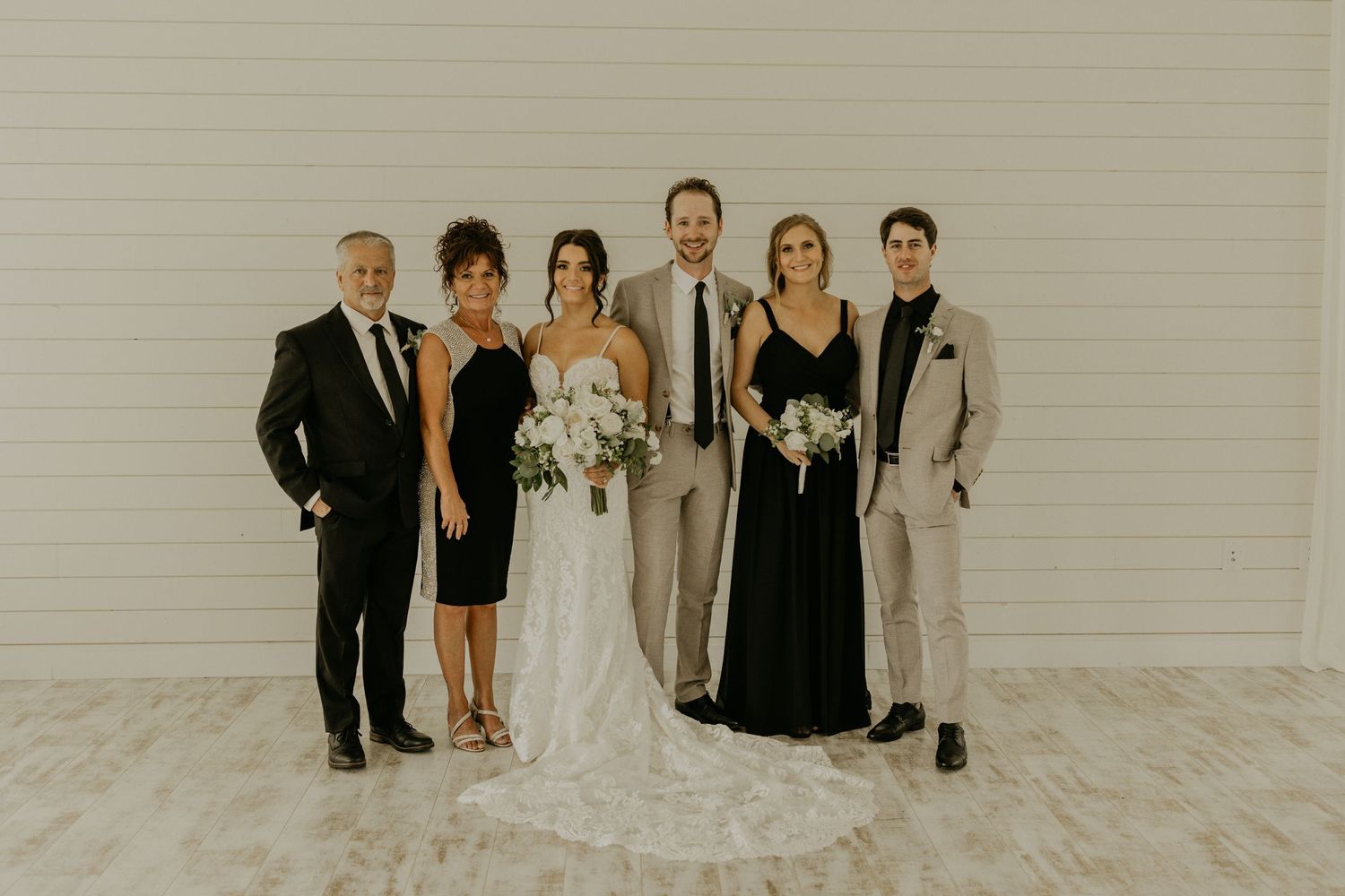 Wedding party poses together against white wall wearing mix of formal black dresses and light suits.