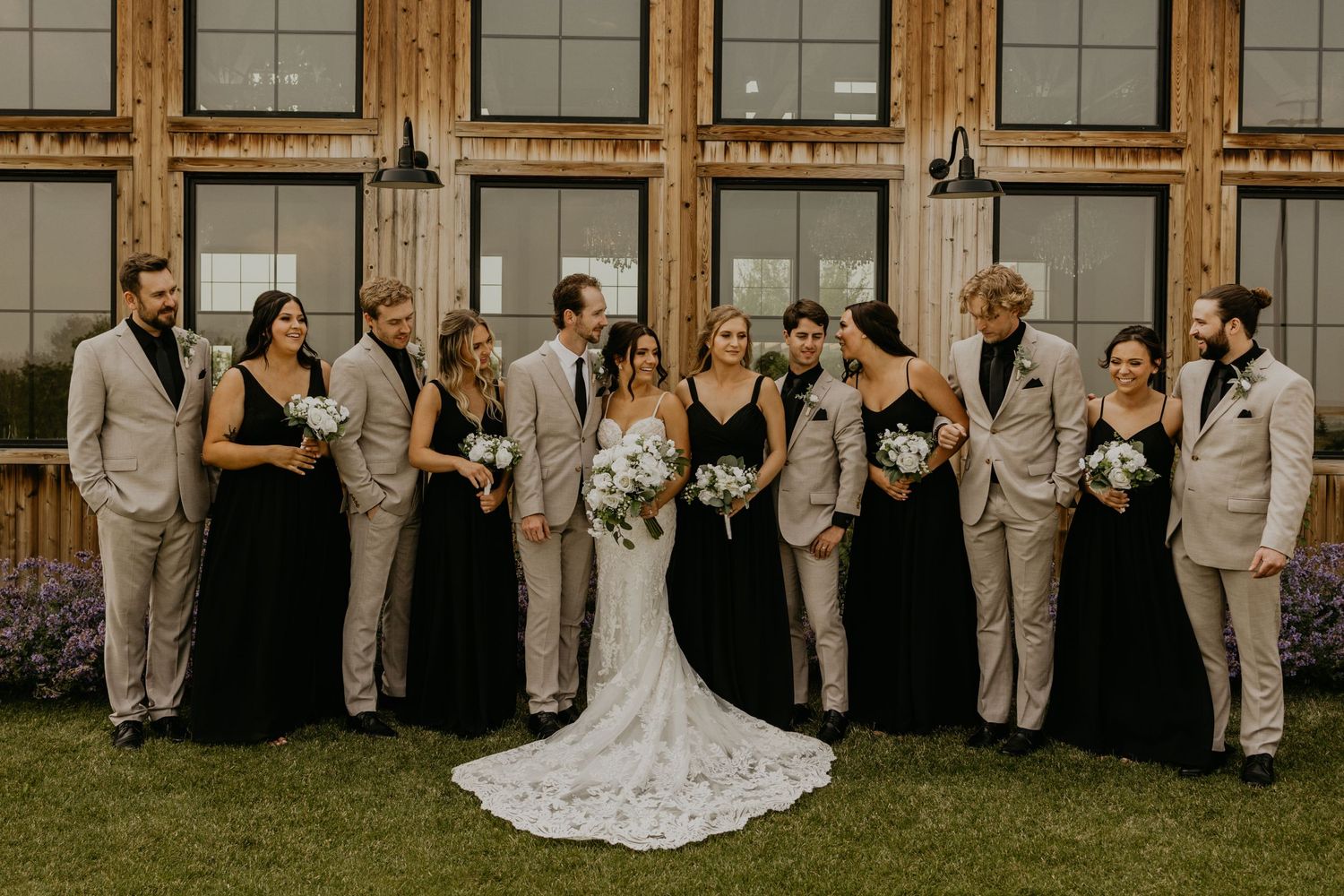 Wedding party laughs and poses together in front of a wooden paneled building.
