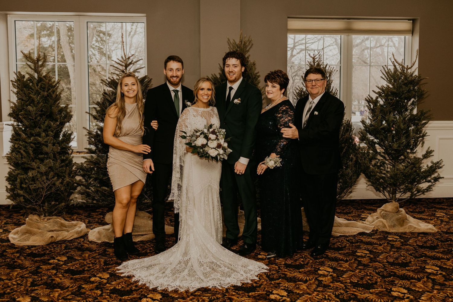 Wedding party poses for formal photos against a backdrop of winter evergreen trees indoors.