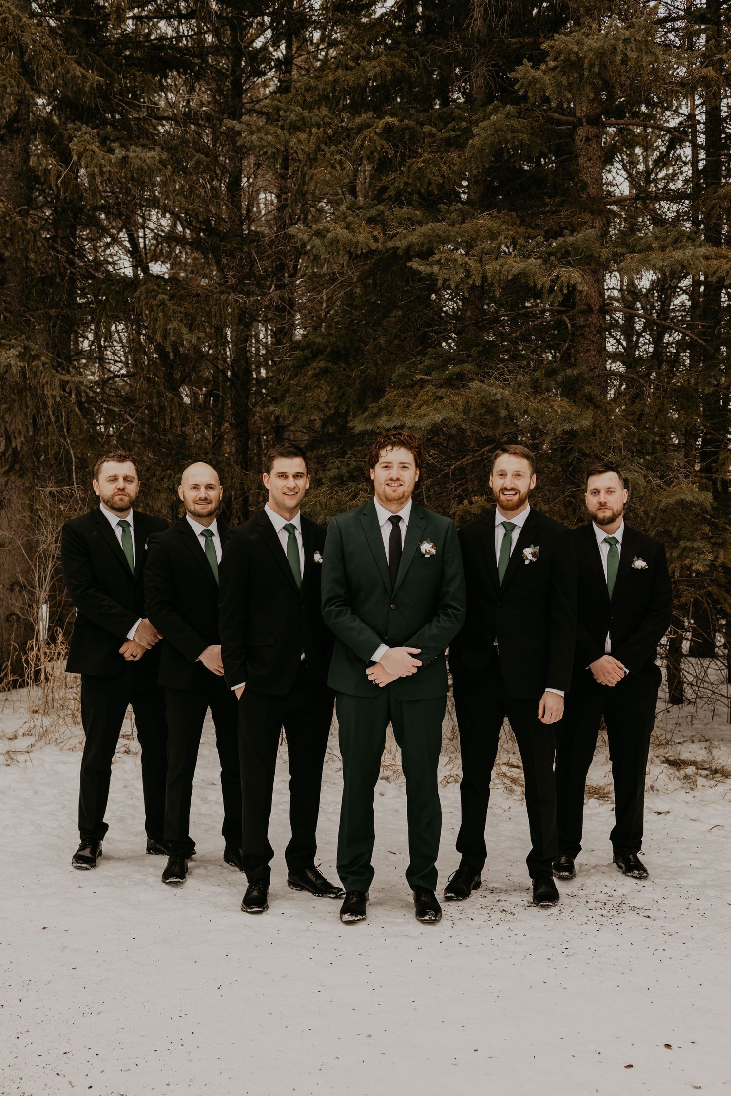 Formal group photo of groomsmen in black suits standing in snow with pine trees behind them.