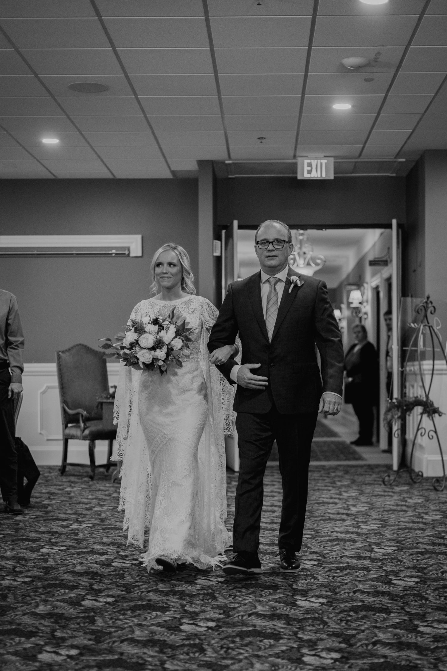 A couple walk arm in arm down a carpeted indoor corridor during a wedding ceremony.