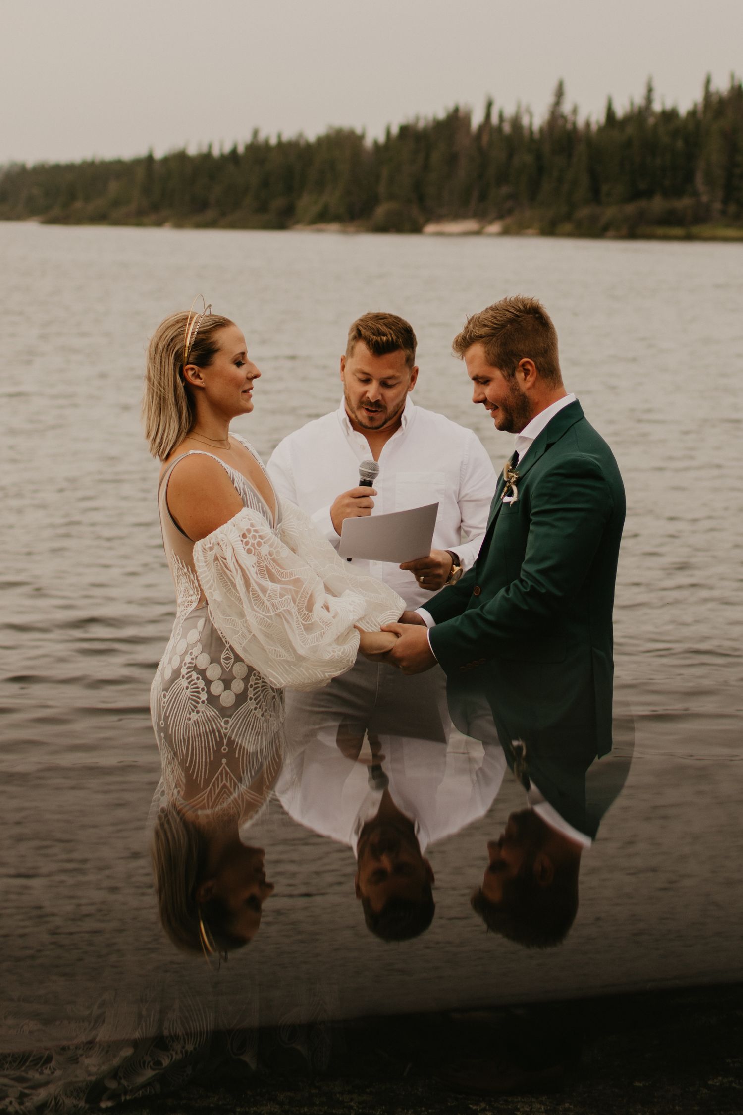 A waterfront wedding ceremony with reflections in the lake and pine trees lining the shore.