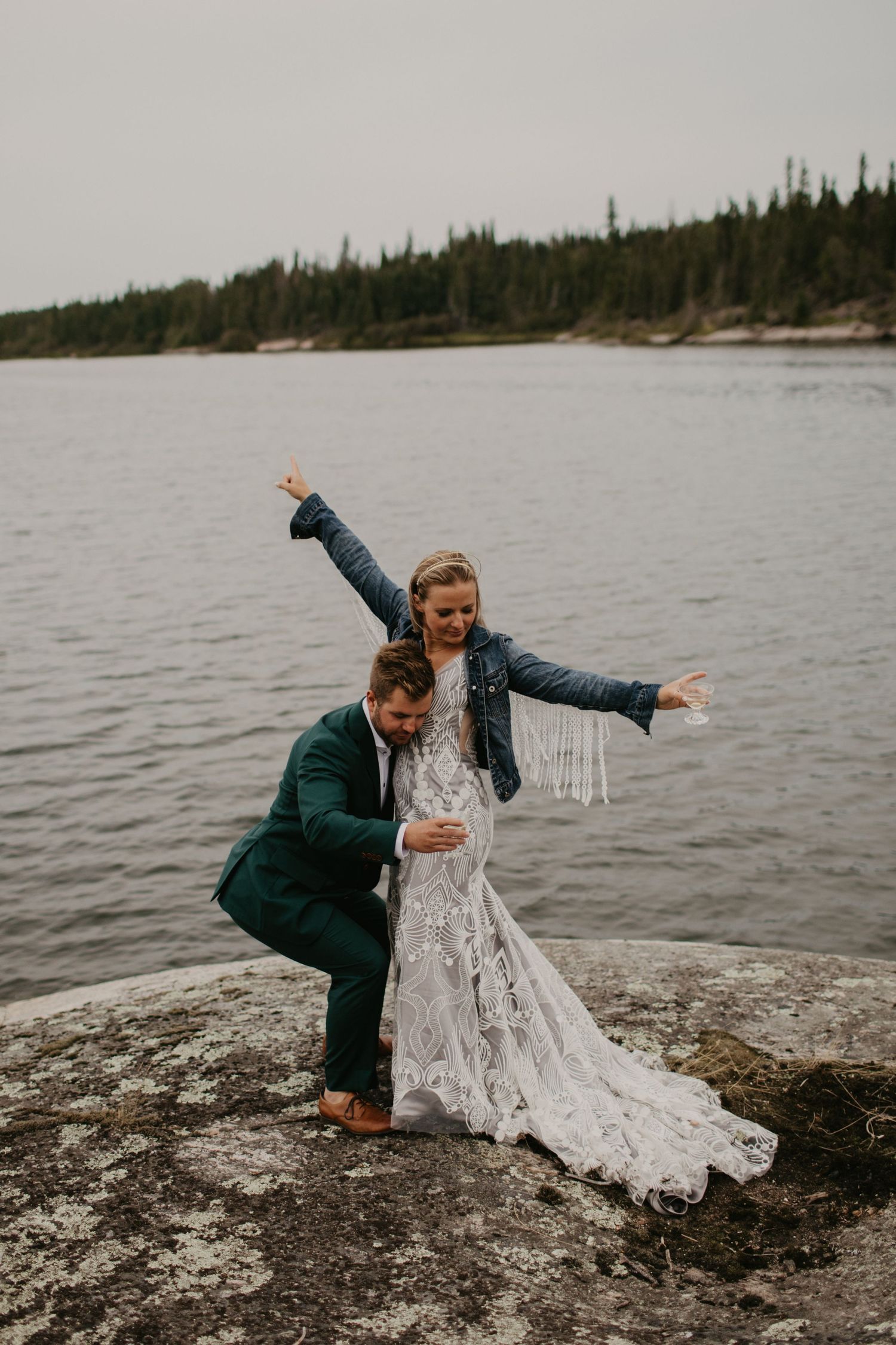 A celebratory moment on a rocky lakeside with a couple in wedding attire and denim jacket.